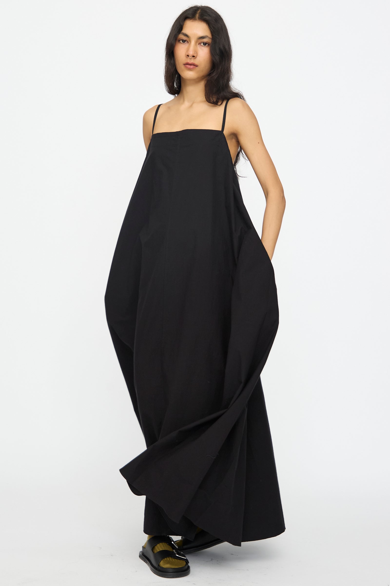A person wears the Studio Nicholson Achille Gathered Dress in Black, a loose cotton maxi with thin straps and a square neckline, standing against a plain white background.