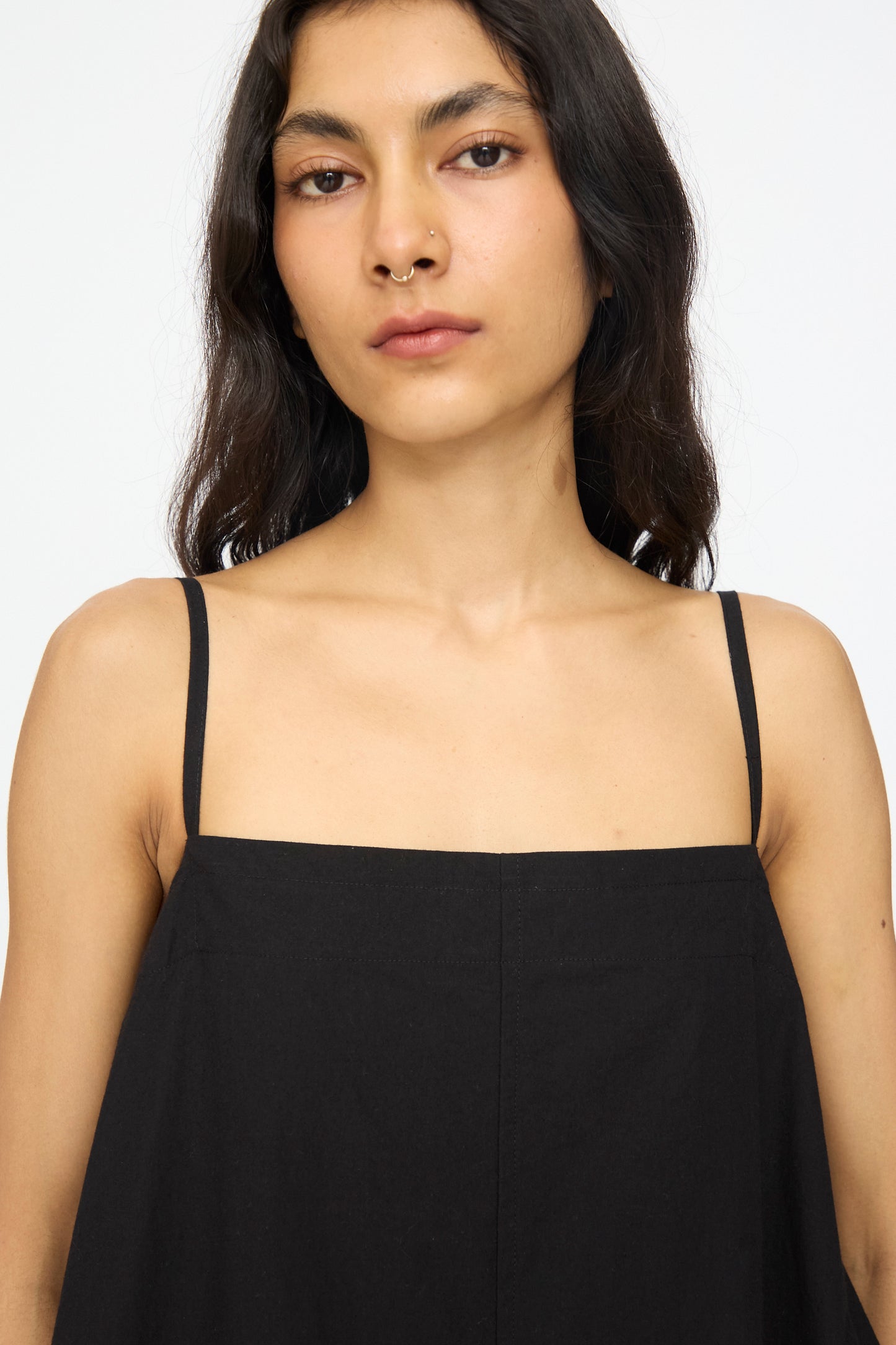 A person with long dark hair, wearing the Studio Nicholson Achille Gathered Dress in Black with a square neckline, faces the camera against a plain white background.
