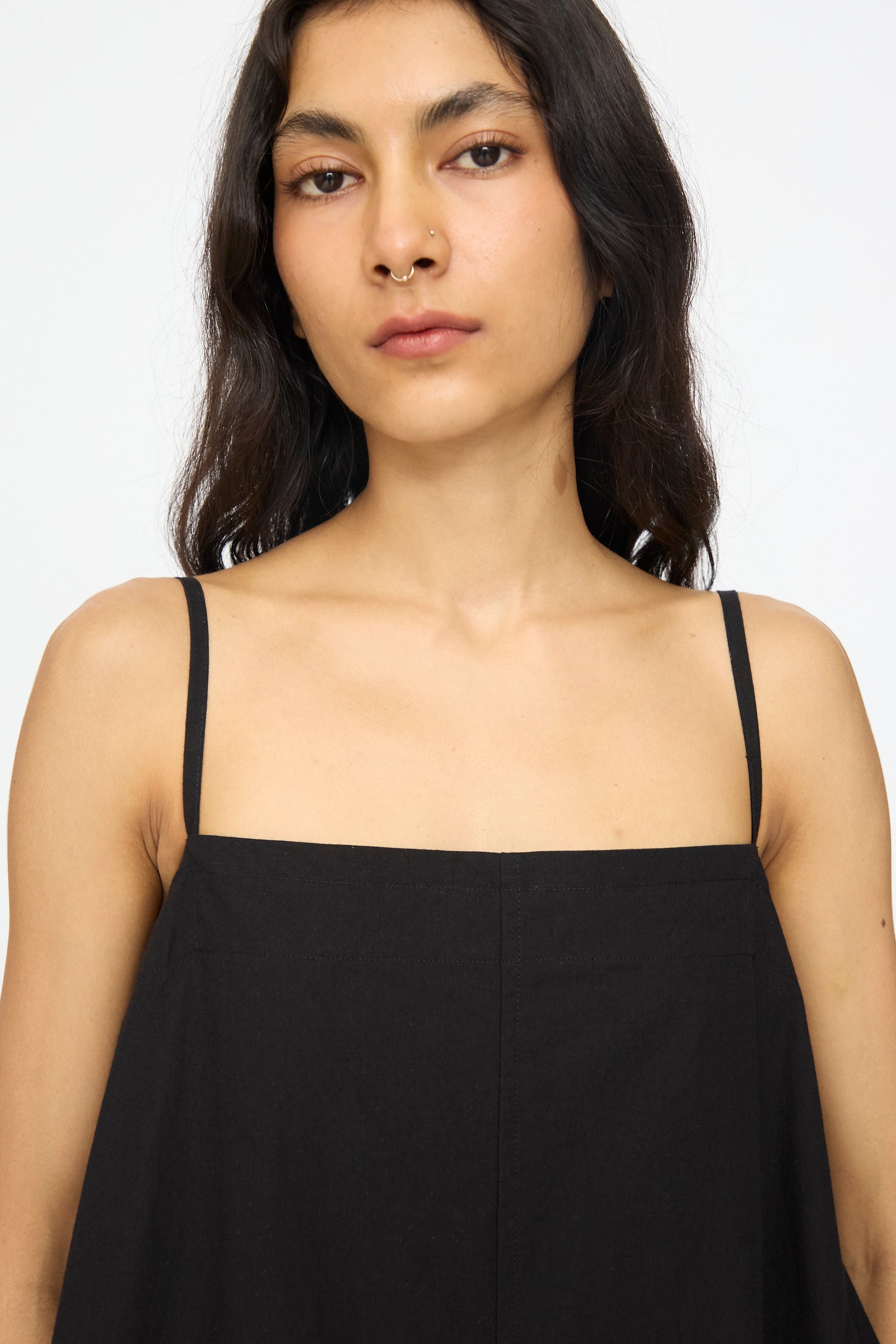A person with long dark hair, wearing the Studio Nicholson Achille Gathered Dress in Black with a square neckline, faces the camera against a plain white background.