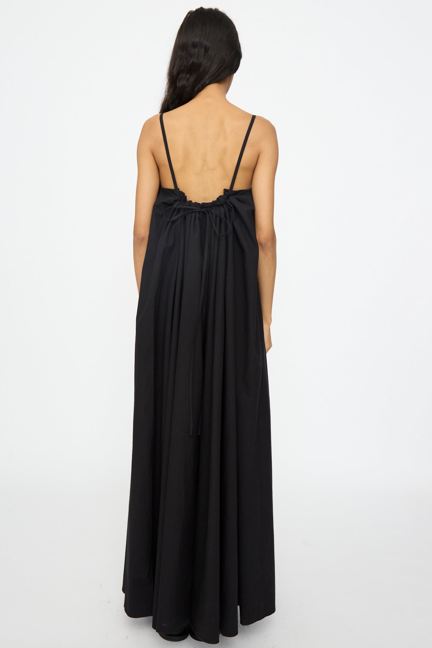 A person with long dark hair, seen from behind, wears the Studio Nicholson Achille Gathered Dress in Black—a sleeveless cotton maxi dress featuring thin straps and an open back.