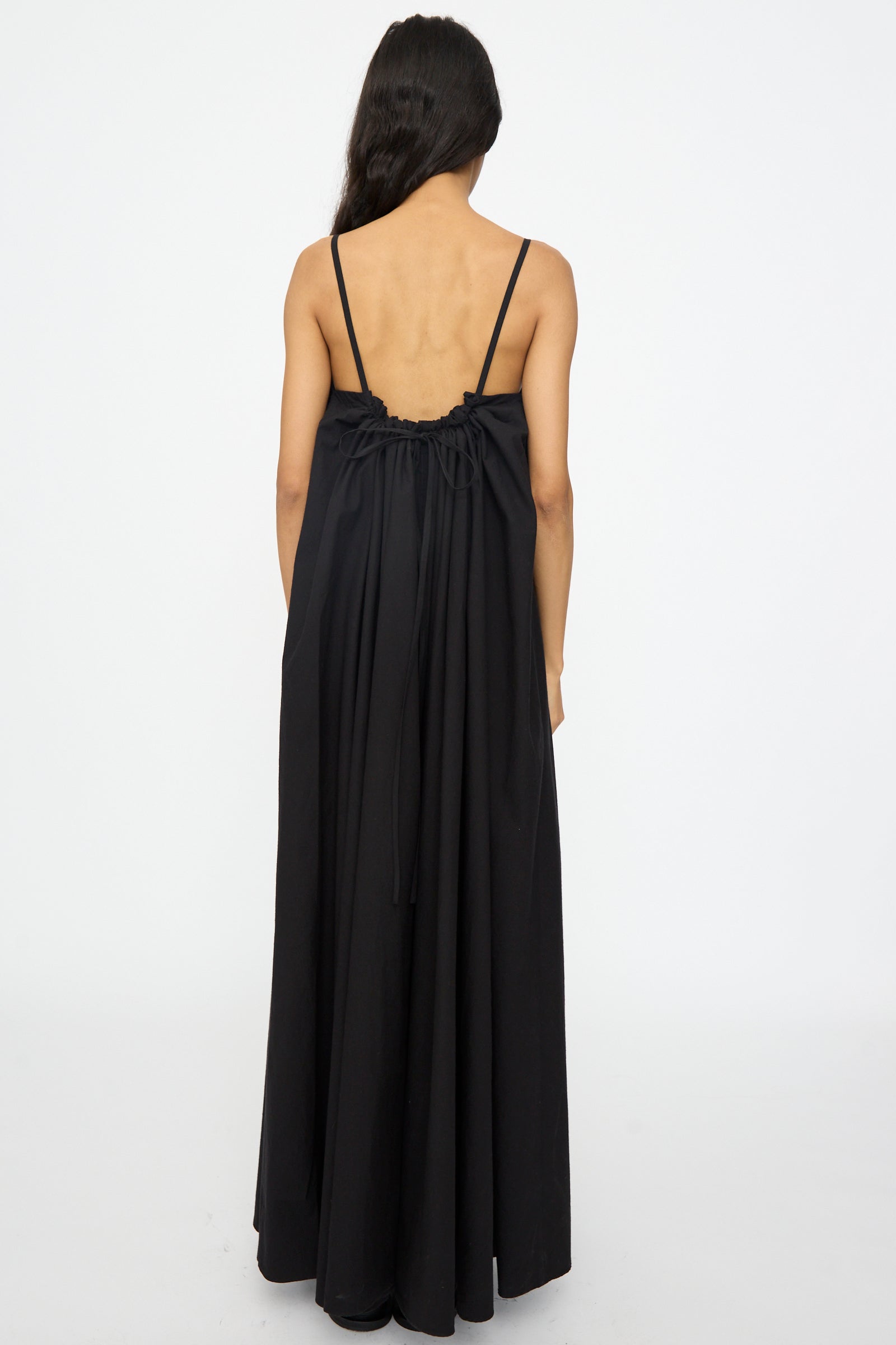 A person with long dark hair, seen from behind, wears the Studio Nicholson Achille Gathered Dress in Black—a sleeveless cotton maxi dress featuring thin straps and an open back.