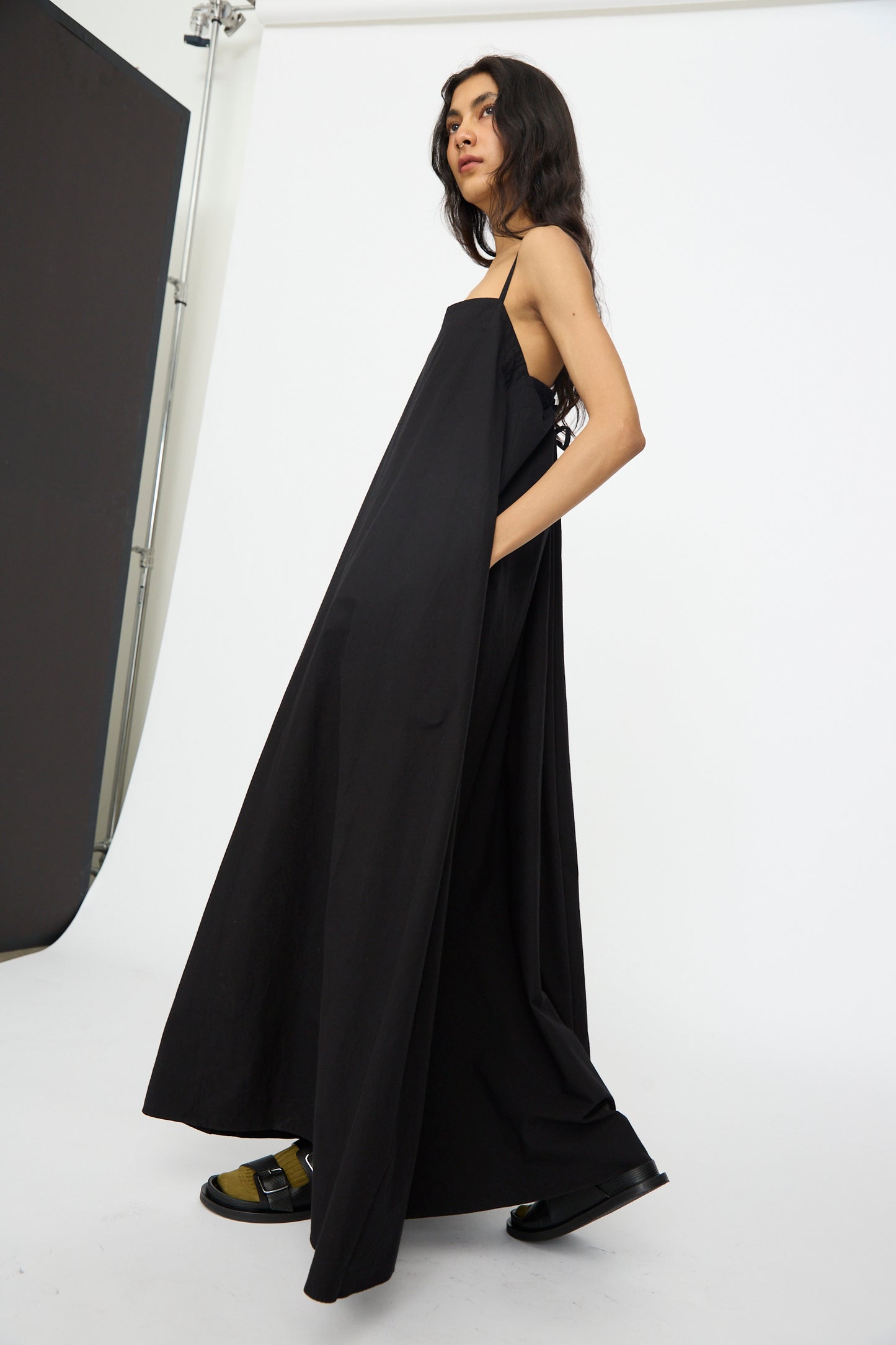 A person stands in a studio with a white background, wearing the Studio Nicholson Achille Gathered Dress in Black and sandals.