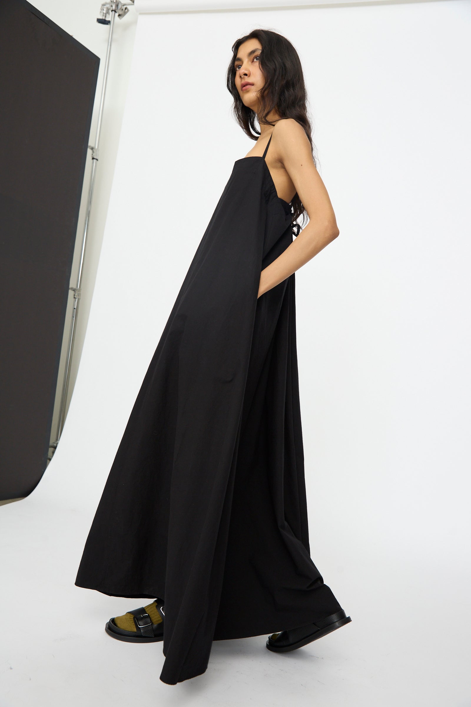 A person stands in a studio with a white background, wearing the Studio Nicholson Achille Gathered Dress in Black and sandals.