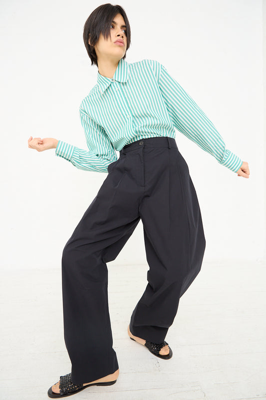 A person in Studio Nicholson's Acuna Double Pleat Front Pant in Darkest Navy and a striped green and white cotton shirt is posing with one hand on their hip and the other arm extended, looking off to the side.