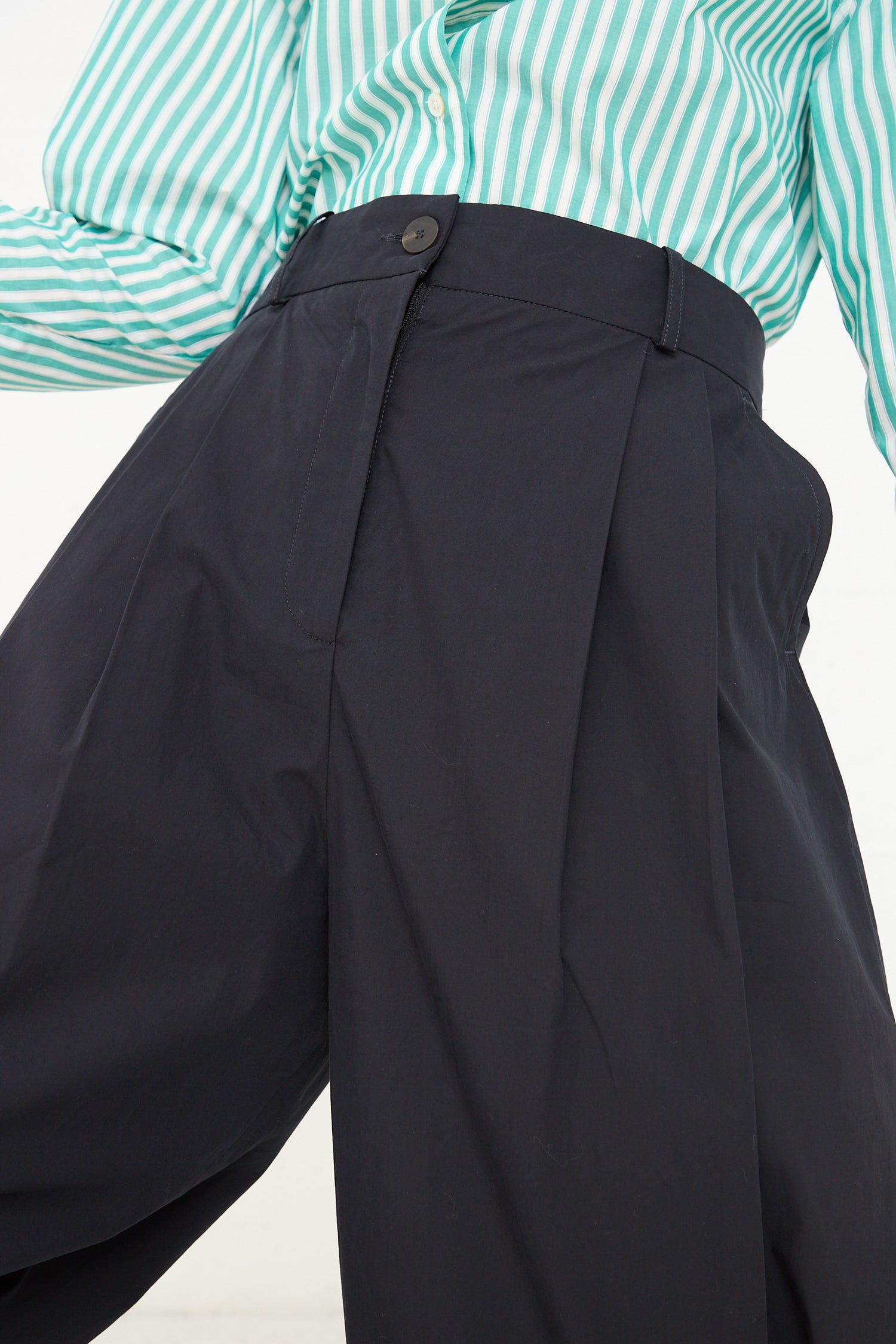 A close-up photo focusing on a person's midsection, featuring a green and white striped cotton shirt tucked into Studio Nicholson's Acuna Double Pleat Front Pant in Darkest Navy with a single visible button.