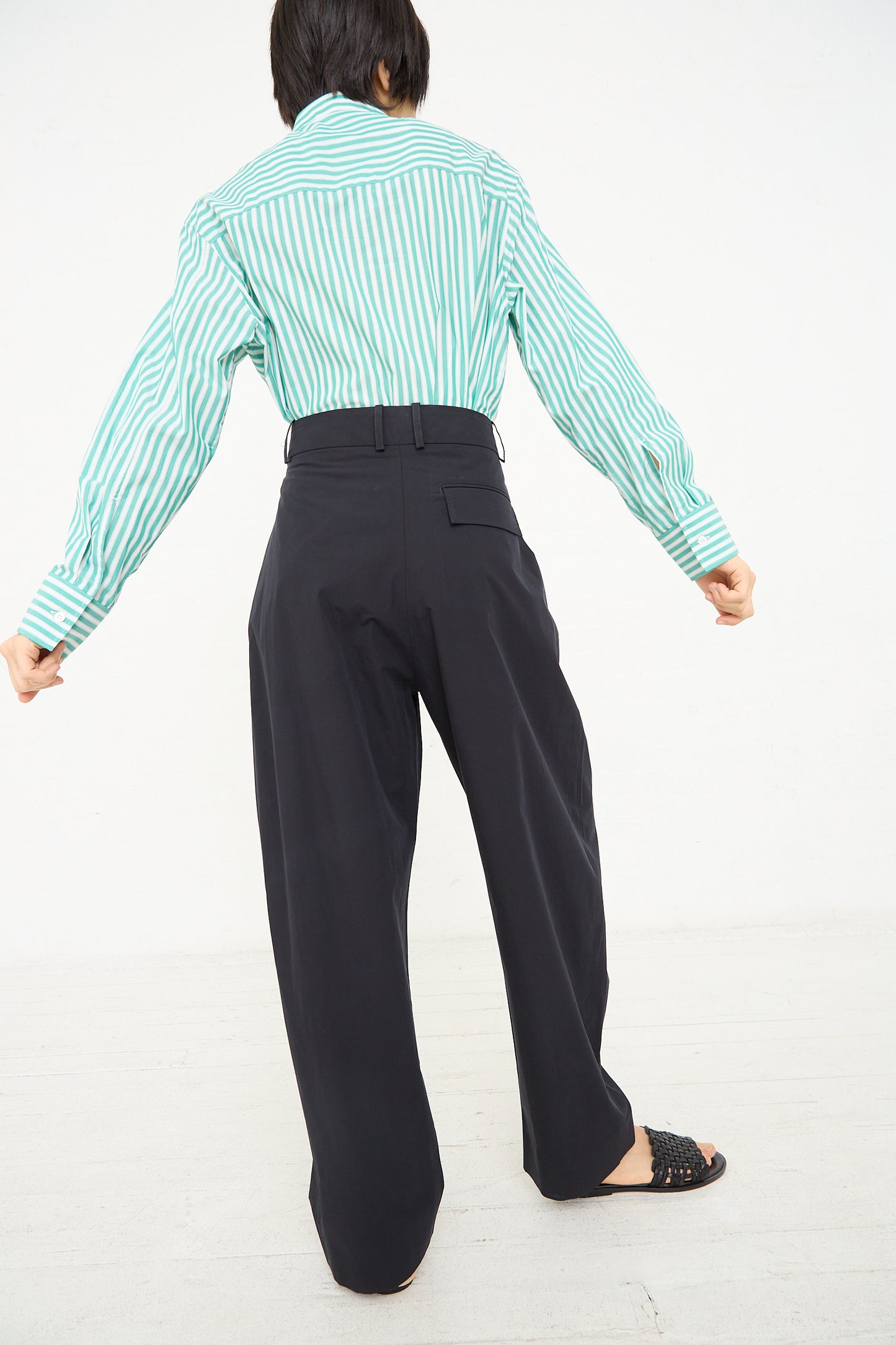 A person facing away from the camera wearing a green and white striped shirt tucked into Studio Nicholson's Acuna Double Pleat Front Pant in Darkest Navy, paired with black slip-on shoes.