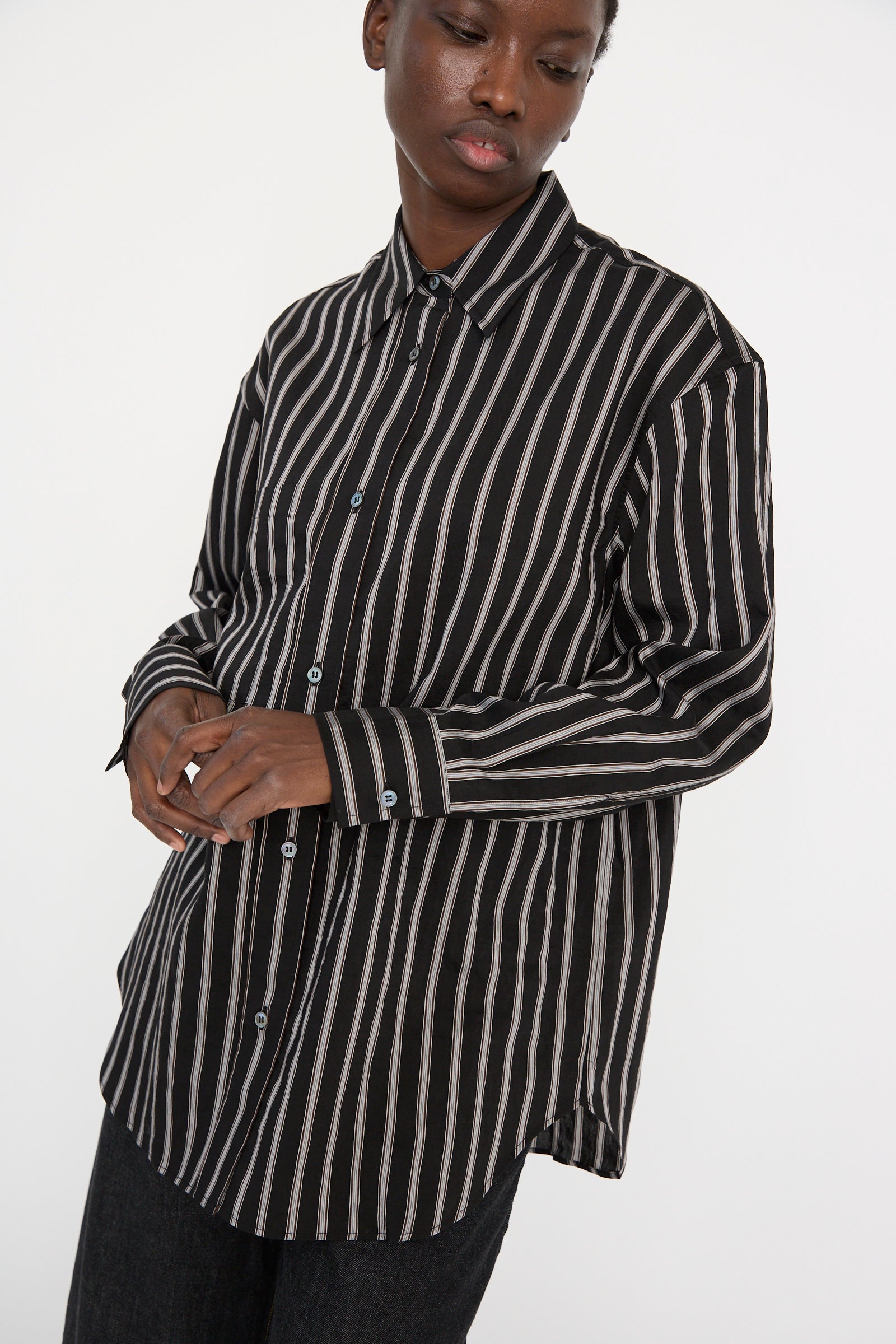 A person is wearing a Studio Nicholson Archive Modal Stripe Leto Shirt in Darkest Navy, looking down with hands clasped against a white backdrop. 