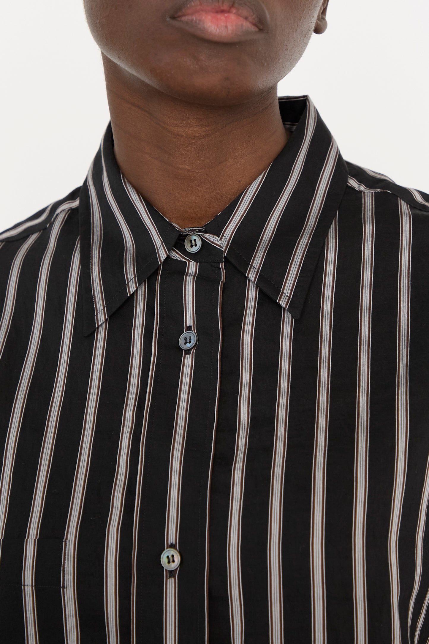 A close-up image of a person wearing the Archive Modal Stripe Leto Shirt in Darkest Navy by Studio Nicholson, an oversized shirt with vertical pin stripes in black, white, and pink hues, complemented by a smart collar.