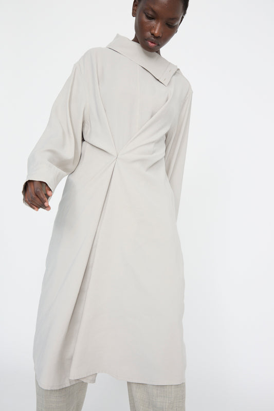 A person looking down is wearing Studio Nicholson's Asymmetric Leat Dress in Gull Grey, set against a plain white background.