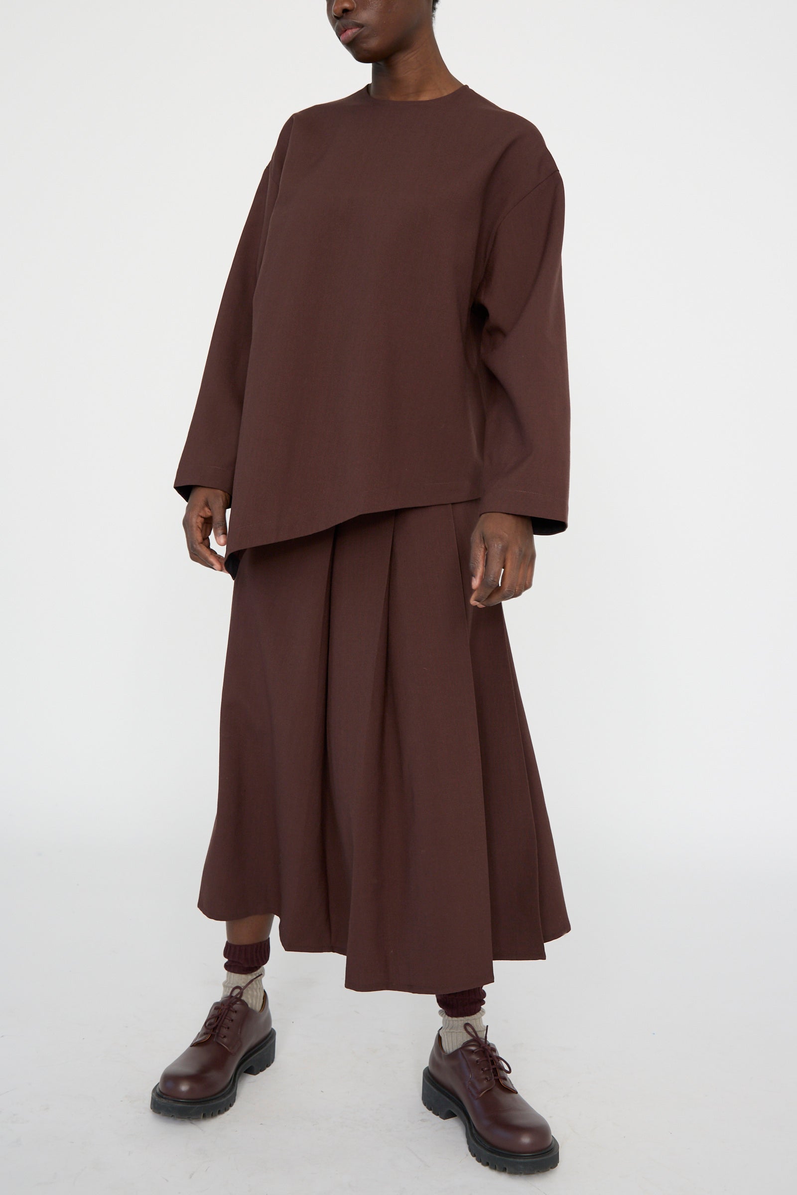 A person stands against a plain background, wearing a brown oversized top with Studio Nicholson's Asymmetric Pleated Fuse Skirt in Conker and matching brown boots.