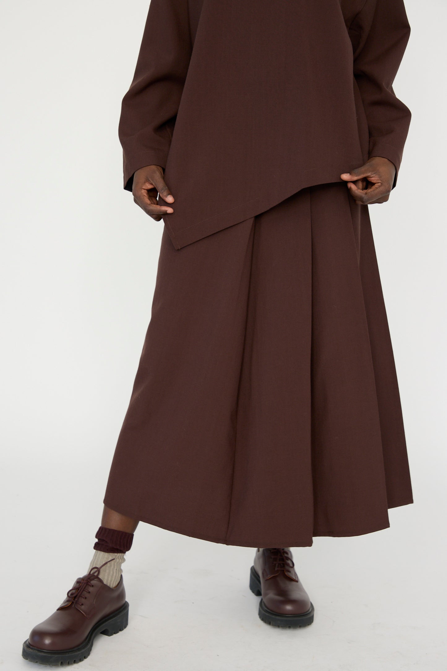 A person wearing Studio Nicholson's Asymmetric Pleated Fuse Skirt in Conker, a matching top, and brown shoes, seen below the shoulders against a white backdrop. 