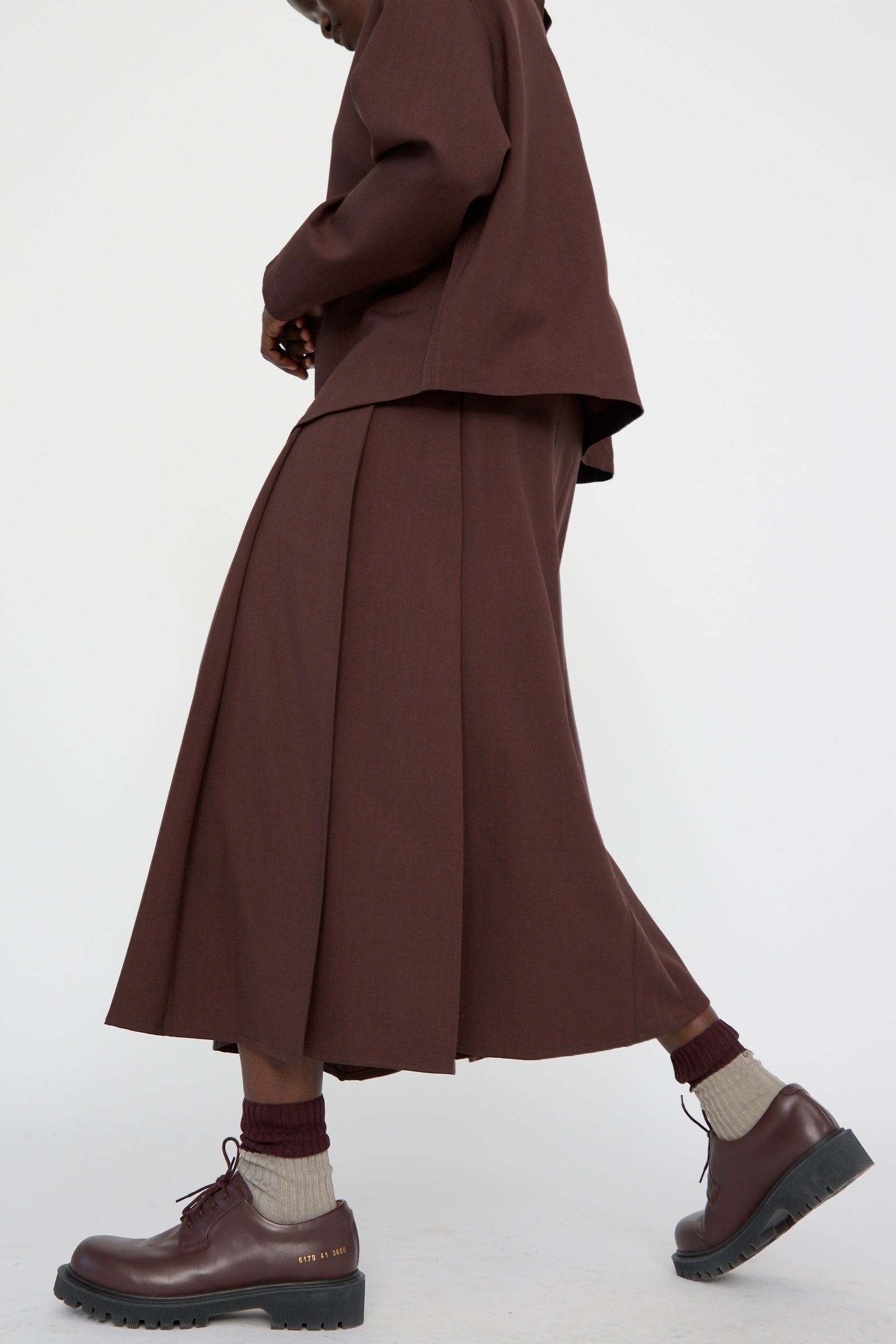 A person wearing a high-waist, brown outfit with an Asymmetric Pleated Fuse Skirt in Conker by Studio Nicholson and brown leather shoes walks to the side against a plain white background.