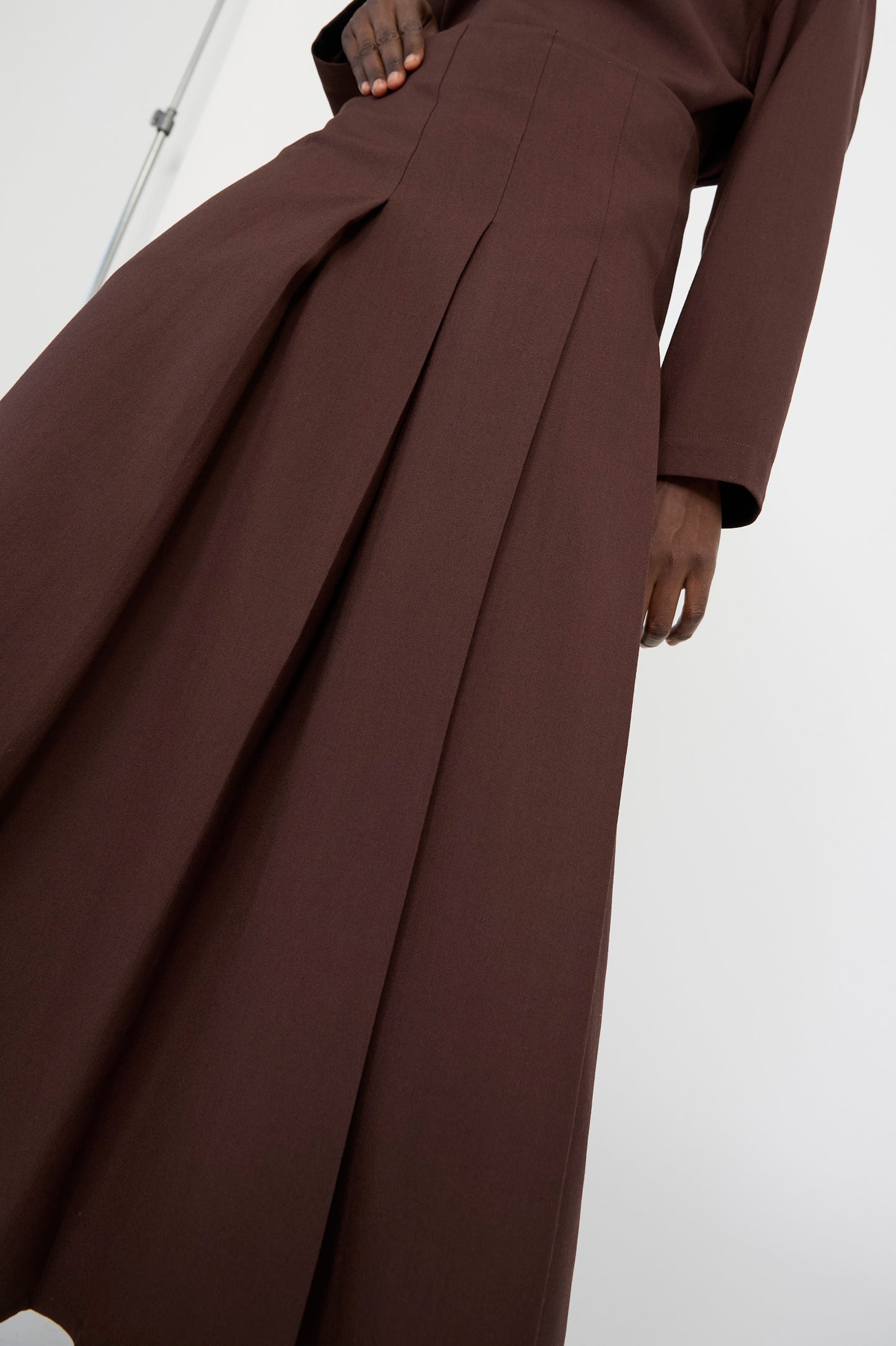 A person in Studio Nicholson's Asymmetric Pleated Fuse Skirt in Conker stands with their hand by their side. The skirt, made of lightweight wool, features long pleats and fine tailoring.