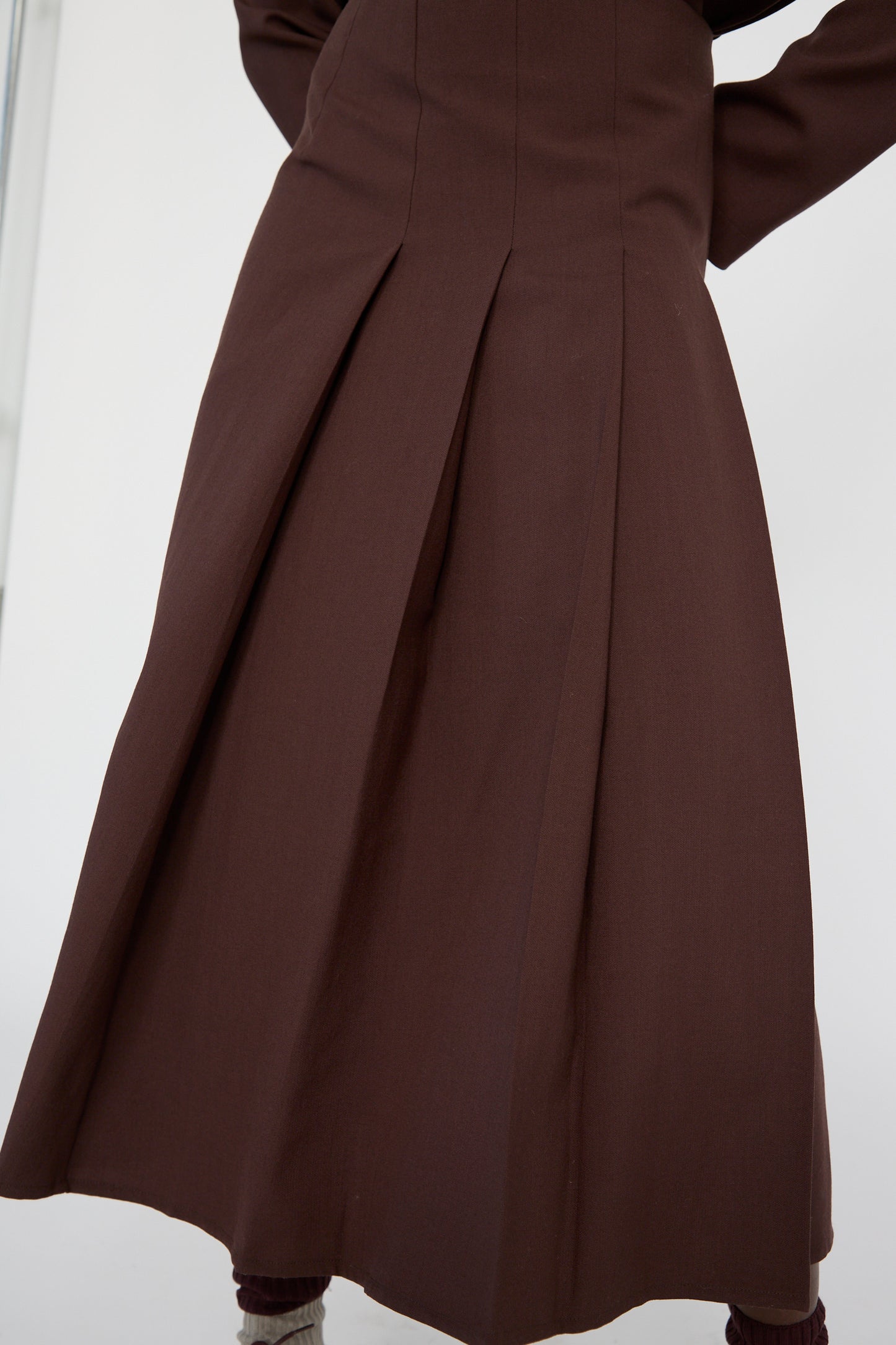 A woman with arms folded wears Studio Nicholson's Asymmetric Pleated Fuse Skirt in Conker, a long dark brown midi skirt offering an elegant high-waisted silhouette.