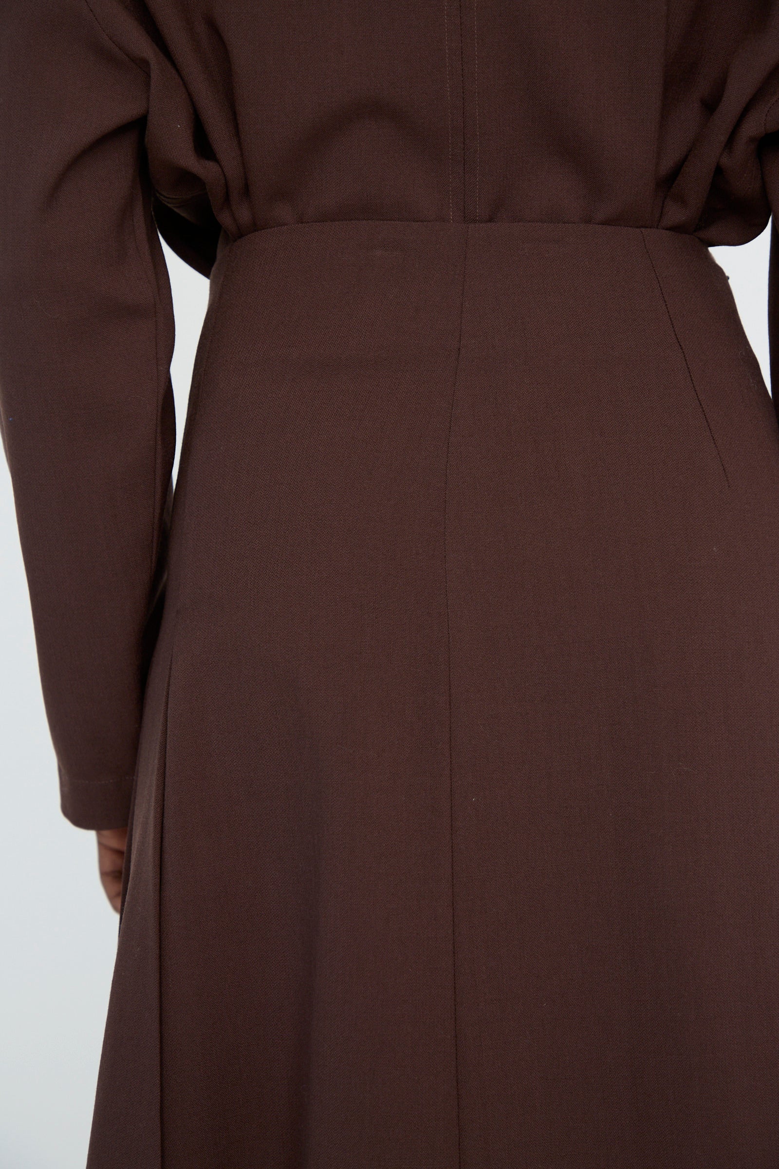 Back view close-up of a person wearing an Asymmetric Pleated Fuse Skirt in Conker by Studio Nicholson, crafted from lightweight wool, showcasing smooth fabric and a seam down the center.