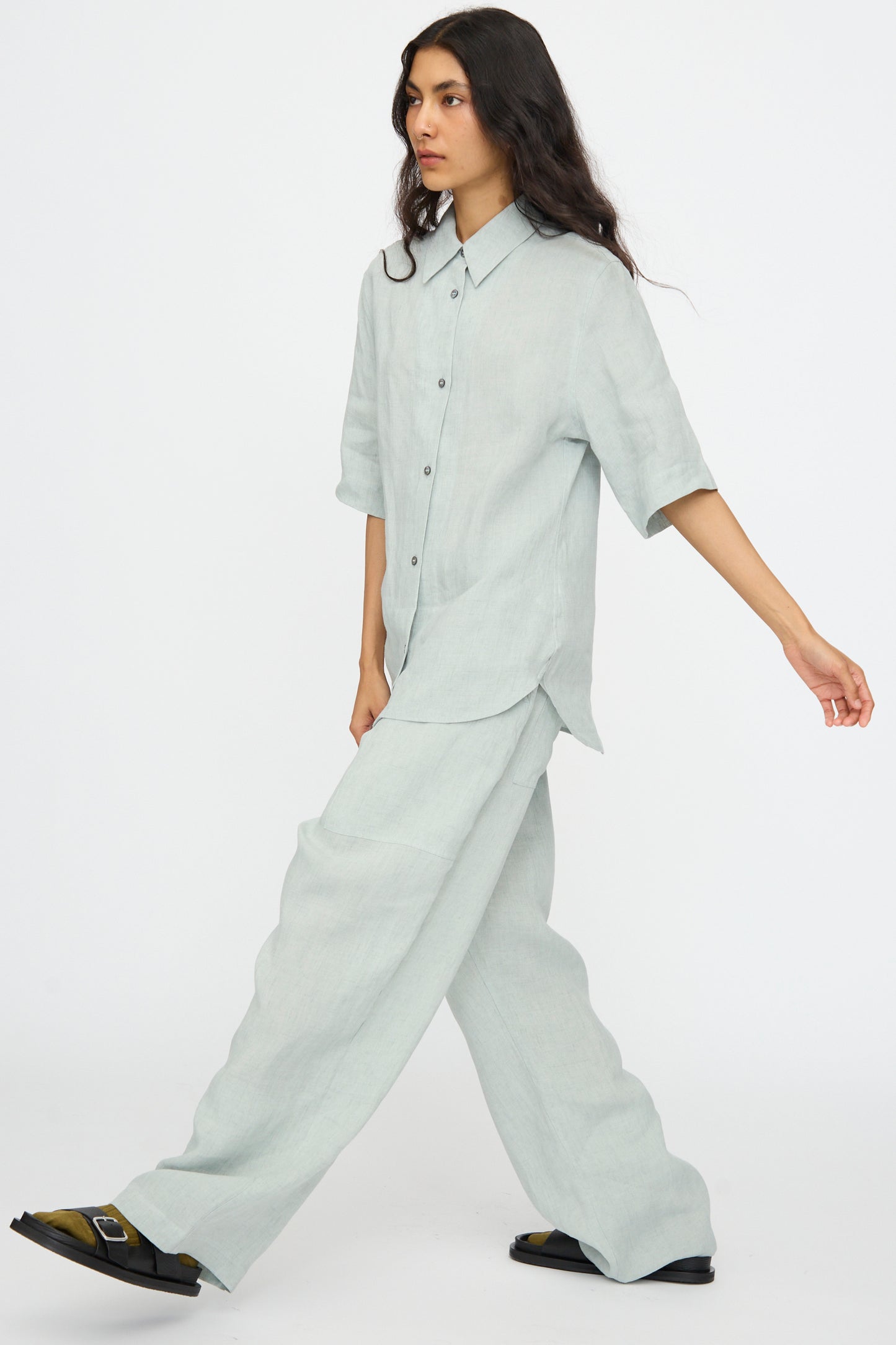 A person wears Studio Nicholson’s Attis Pant in Spearmint, styled with a loose light gray button-up shirt and black sandals, set against a plain white background.