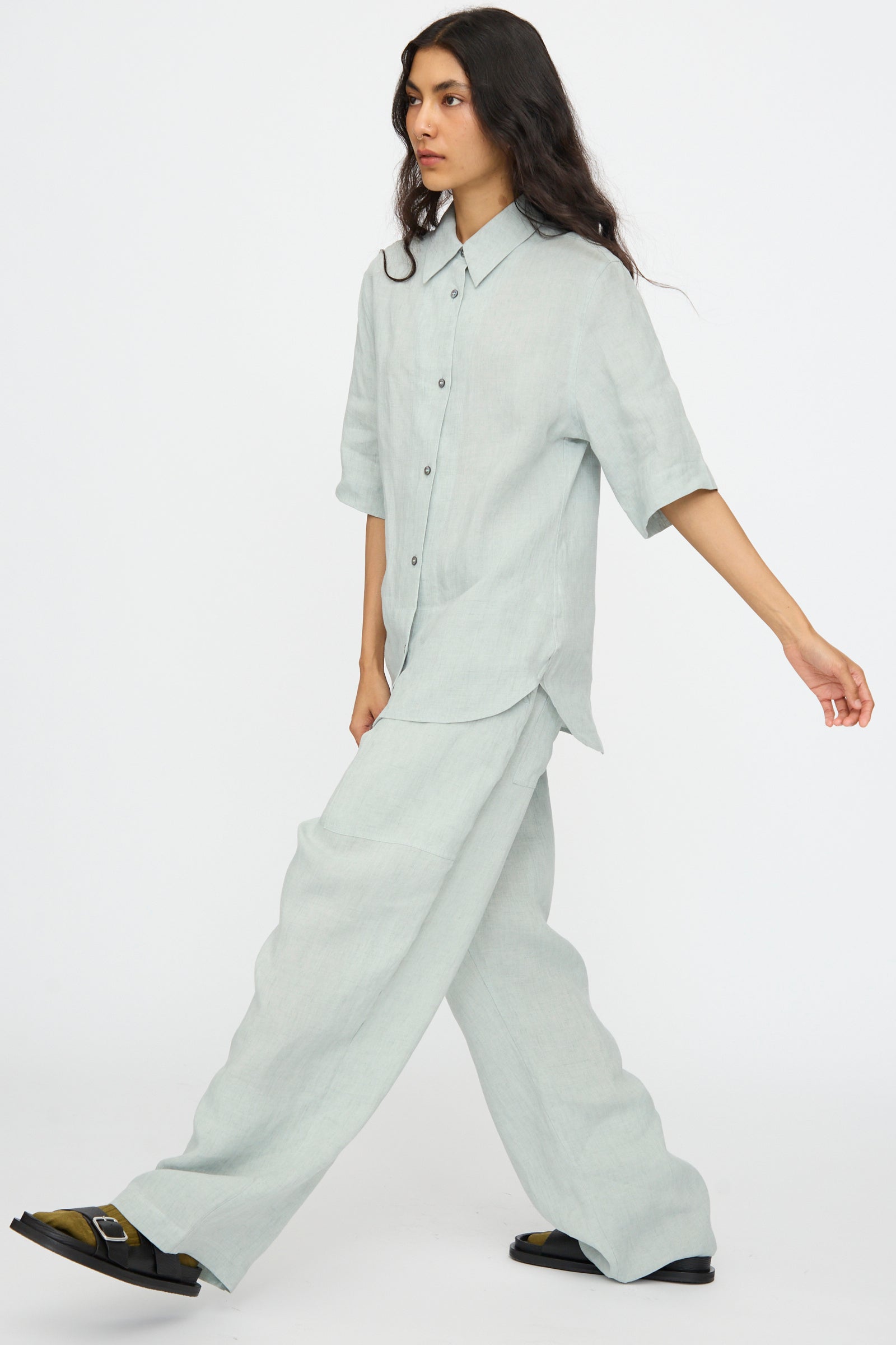 A person wears Studio Nicholson’s Attis Pant in Spearmint, styled with a loose light gray button-up shirt and black sandals, set against a plain white background.