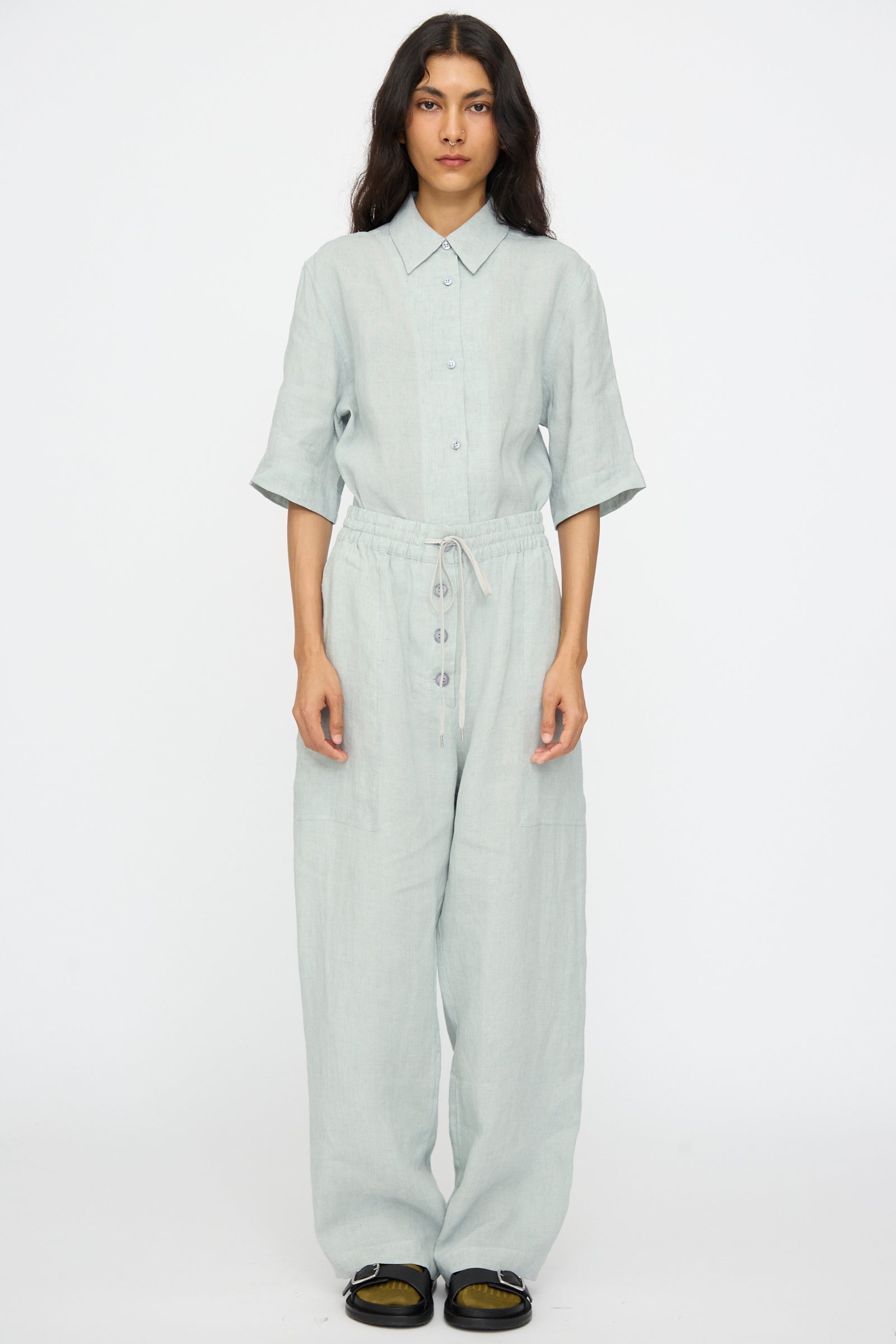 A person stands facing forward in the Studio Nicholson Attis Pant in Spearmint, featuring a wide leg and elastic waistband, paired with a light gray button-up shirt and black shoes, against a plain white background.