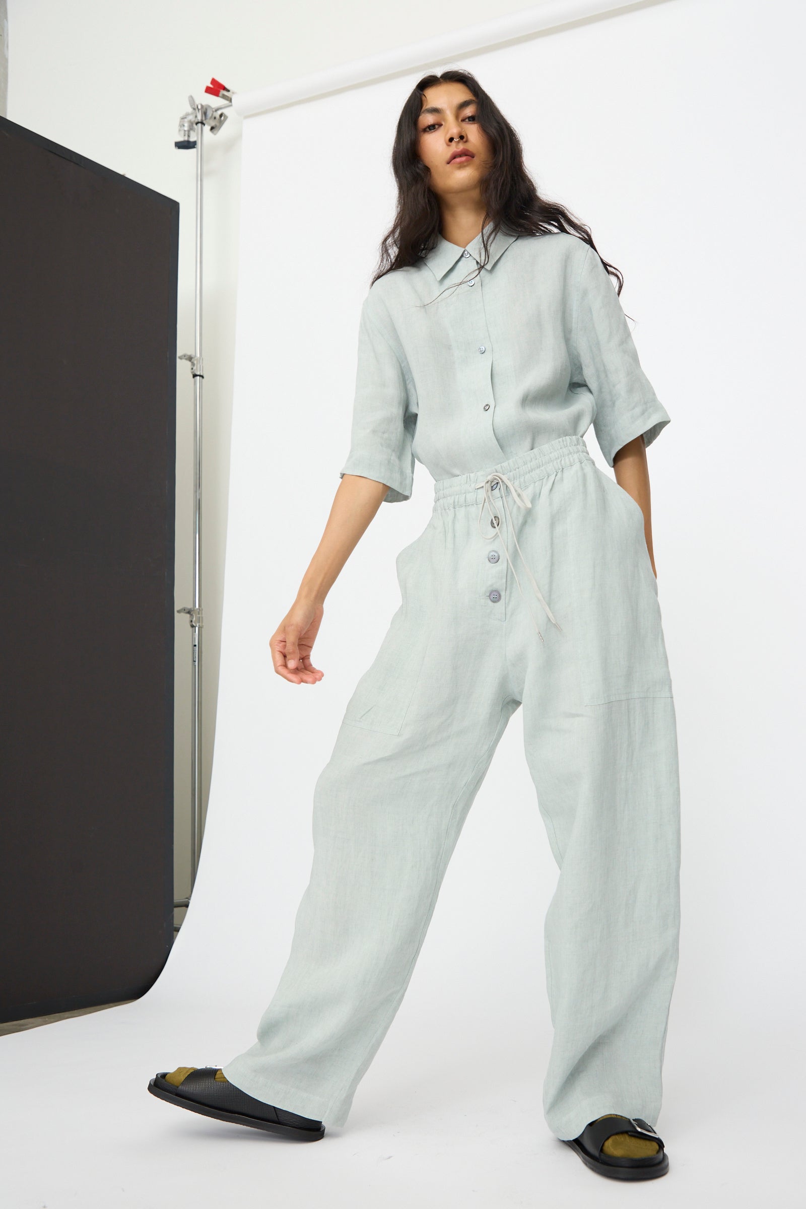 A woman stands before a white backdrop wearing Studio Nicholson's Attis Pant in Spearmint—wide leg linen pants with an elastic waistband—paired with a loose light gray buttoned shirt and black slide sandals.