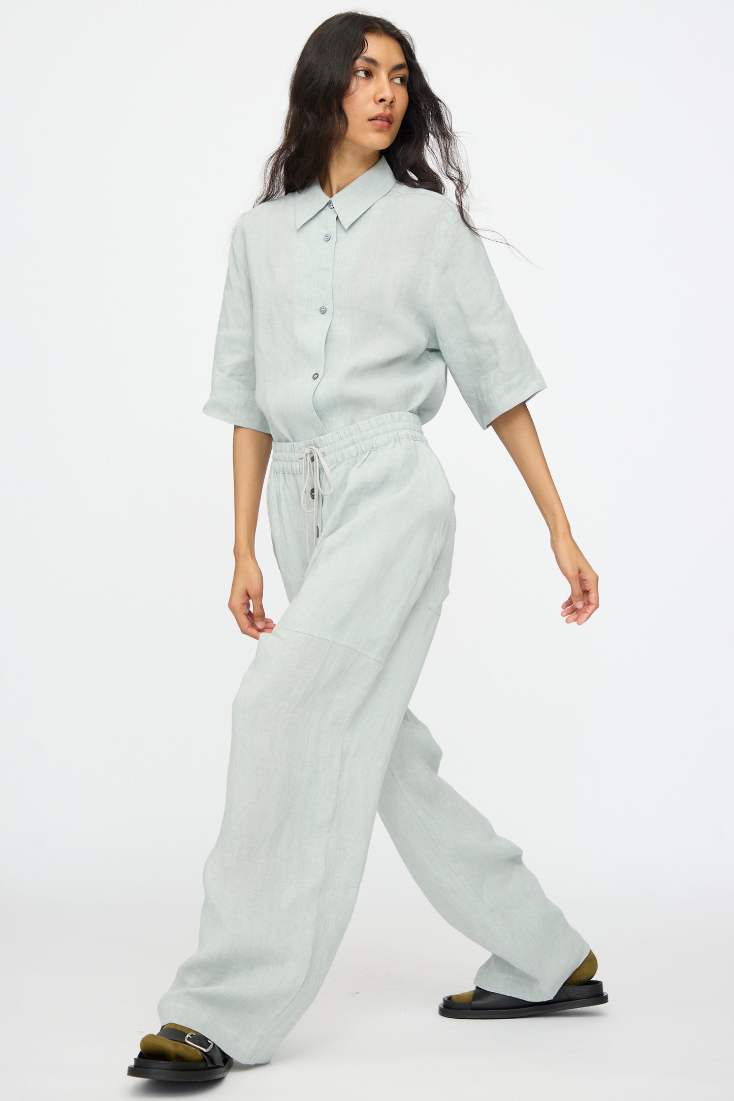 A person walks against a plain white background, wearing the Studio Nicholson Attis Pant in Spearmint—a loose, wide-leg style with an elastic waistband—paired with a light grey button-up shirt.