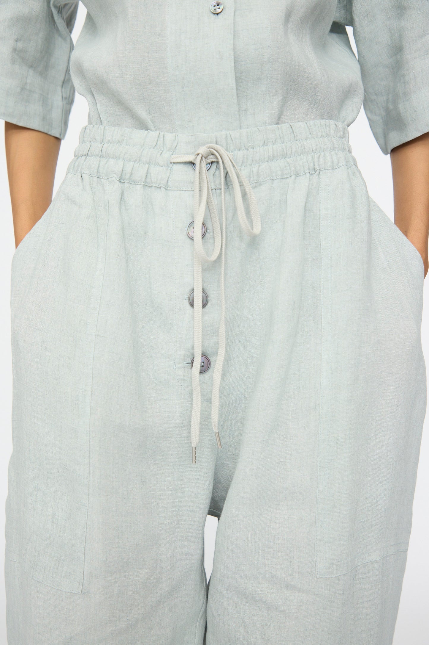 A person wears the Studio Nicholson Attis Pant in Spearmint—light grey, wide-leg linen pants featuring large front pockets, visible buttons, and an elastic waistband—with hands placed in the pockets.