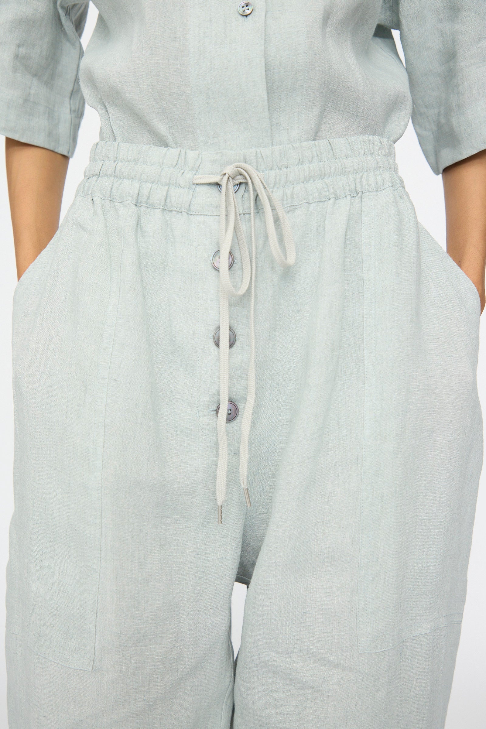 A person wears the Studio Nicholson Attis Pant in Spearmint—light grey, wide-leg linen pants featuring large front pockets, visible buttons, and an elastic waistband—with hands placed in the pockets.