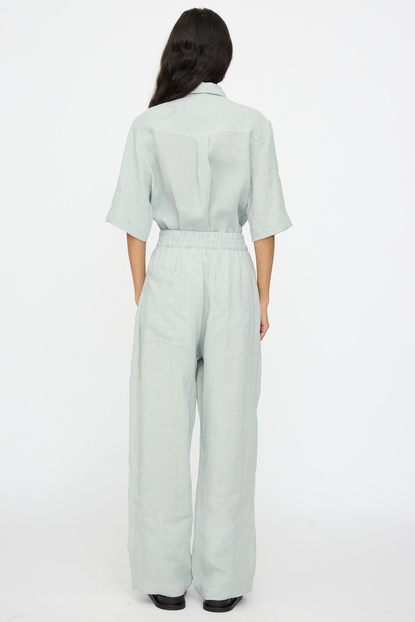 A person stands with their back to the camera, wearing Studio Nicholson’s Attis Pant in Spearmint—light-colored, loose-fitting with an elastic waistband and wide legs—paired with black shoes against a plain white background.