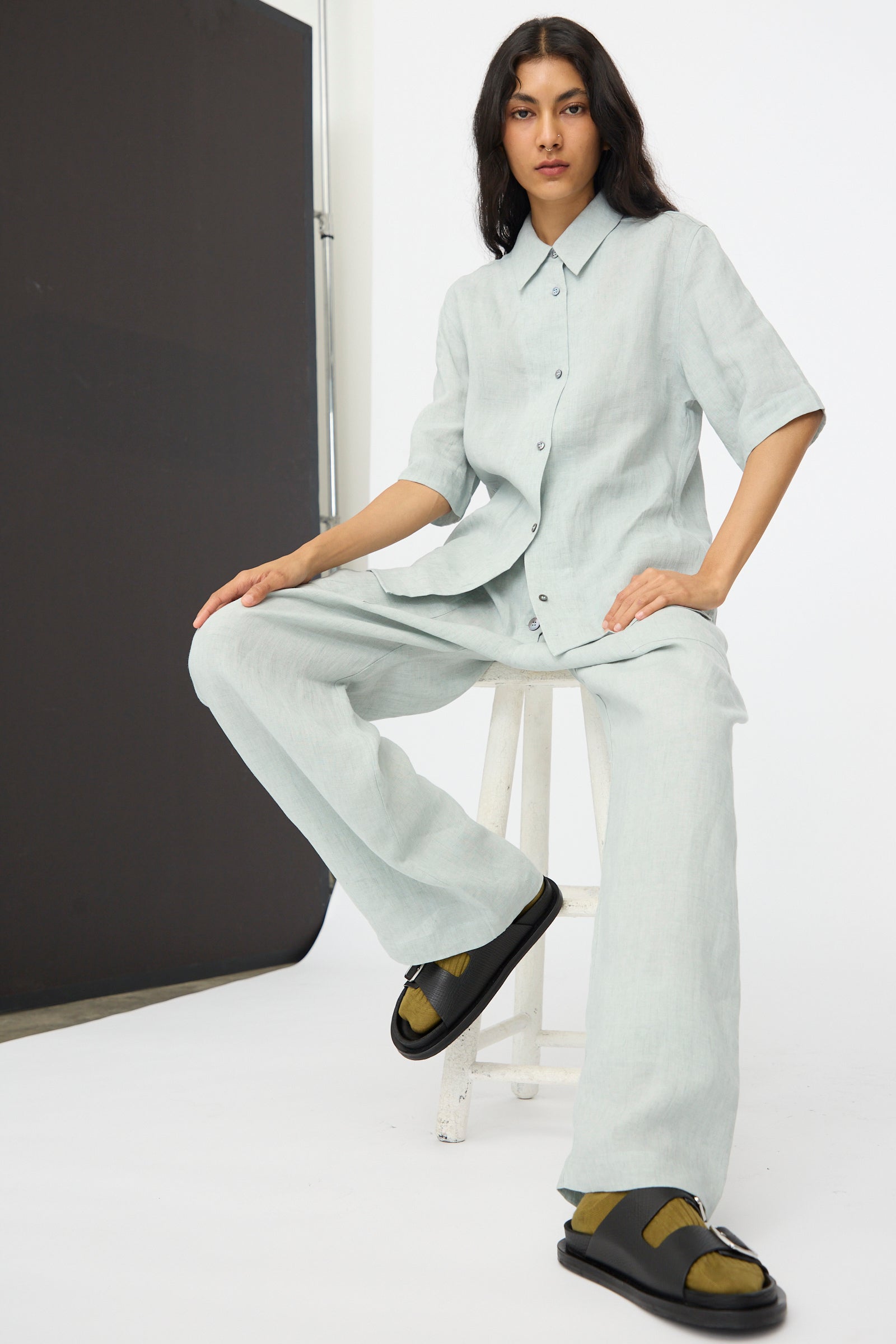 A person with long hair sits on a white stool in a minimalist studio, wearing a light button-up shirt, dark slide sandals, and the Studio Nicholson Attis Pant in Spearmint featuring an elastic waistband and wide-leg linen design.