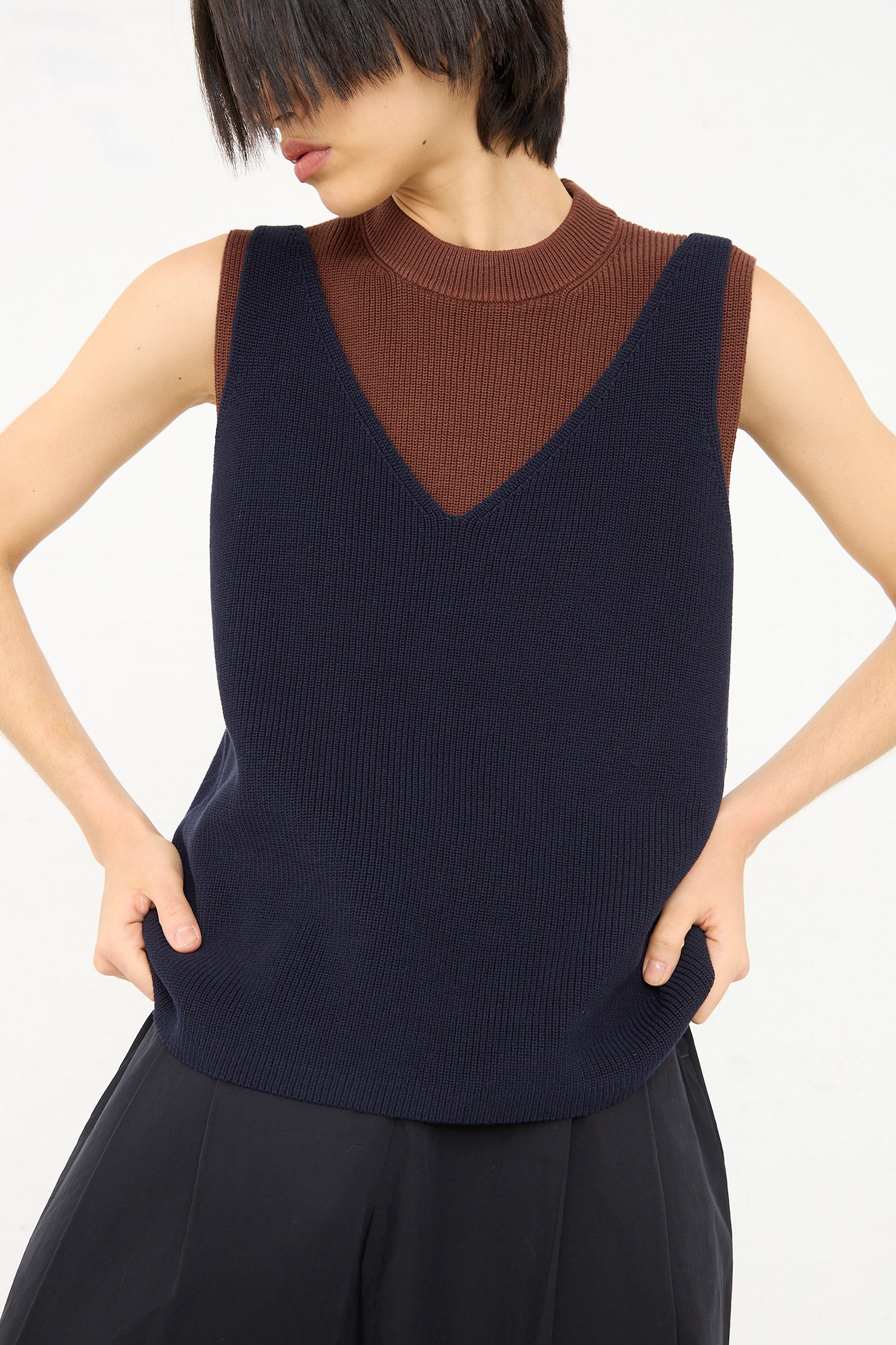 A person wearing a layered outfit with a sleeveless deep V Studio Nicholson Darkest Navy Bara Knit Rib Top over a brown turtleneck, hands casually placed in the pockets of a black garment.