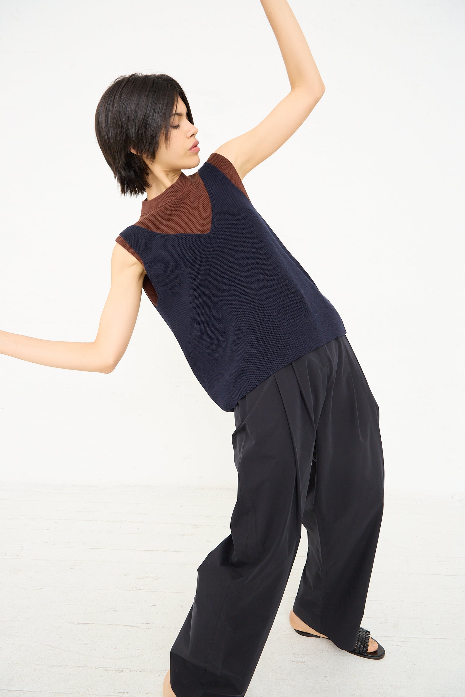 A person with short hair, wearing a Bara Knit Rib Top in Darkest Navy and wide-legged pants from Studio Nicholson, is captured mid-pose against a white background.