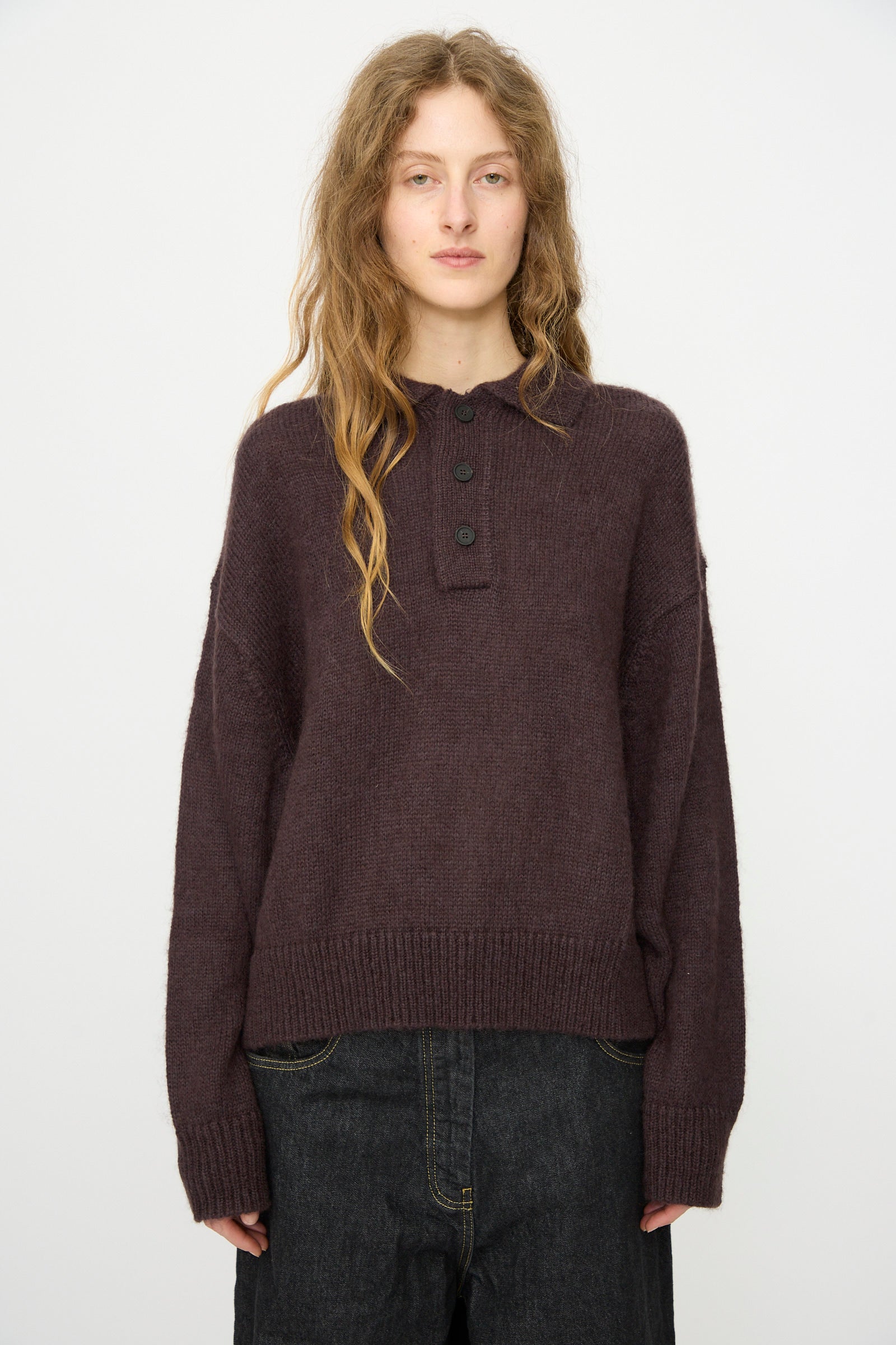 A person with long, wavy hair wears the Studio Nicholson Brushed Mohair Dune Knit Shirt in Espresso and dark jeans, standing against a plain white background.