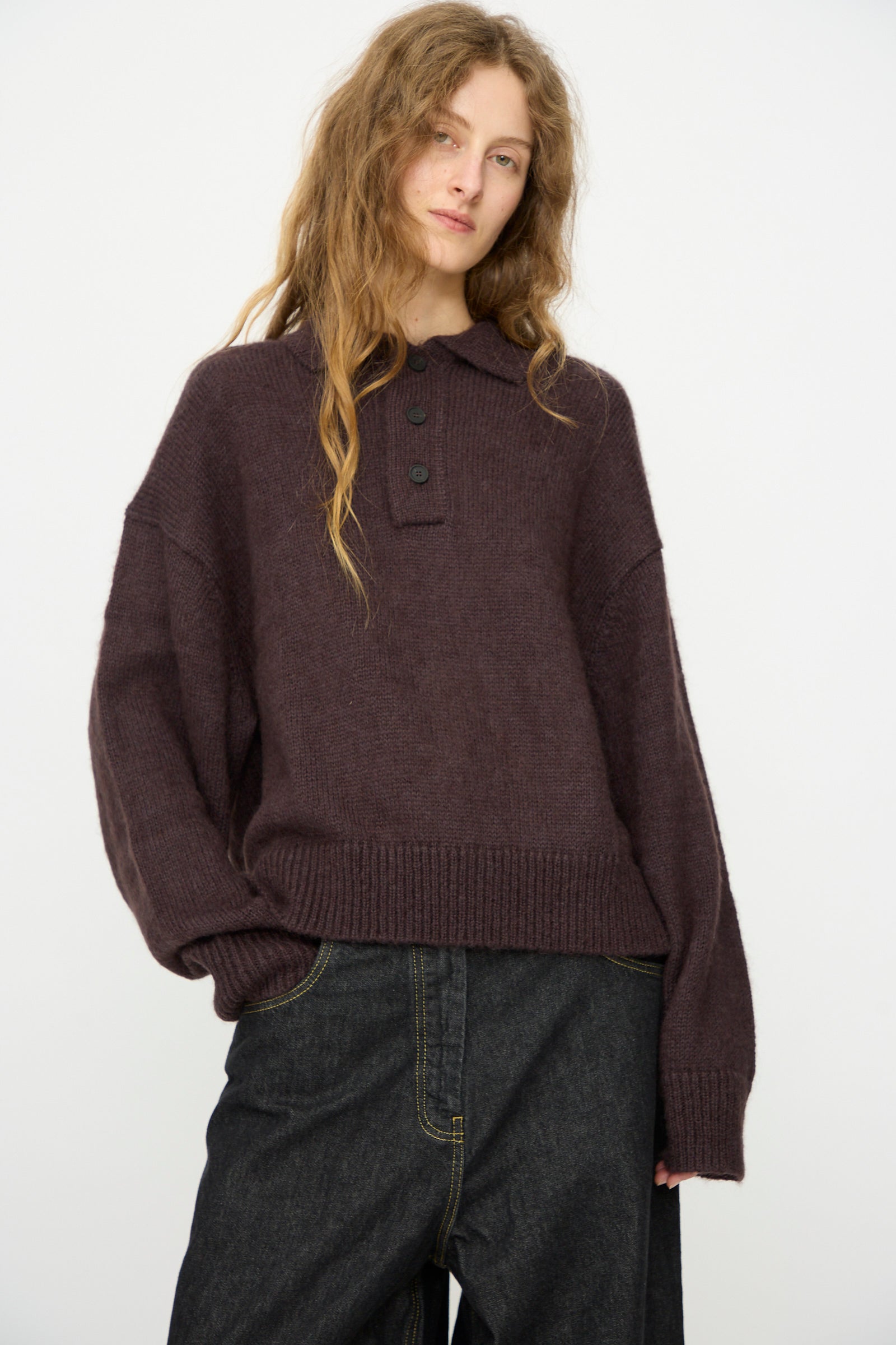 A person with long wavy hair wears the Studio Nicholson Brushed Mohair Dune Knit Shirt in Espresso and dark wide-leg jeans, standing against a plain light background.