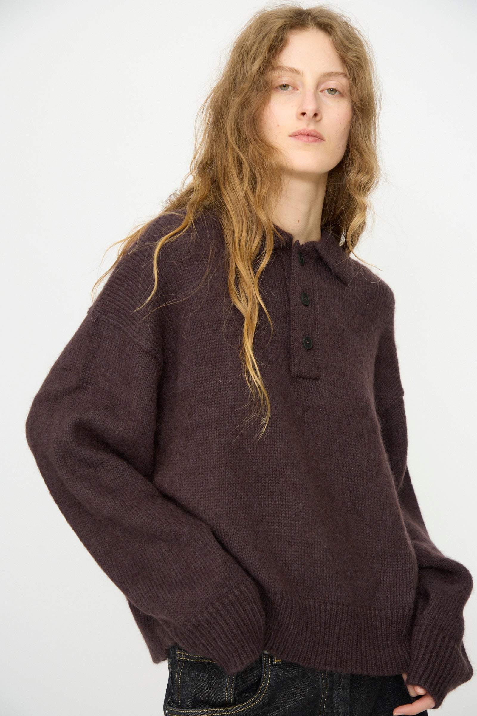 A person with long wavy hair wears the Studio Nicholson Brushed Mohair Dune Knit Shirt in Espresso, featuring a collar and buttons, styled with dark jeans and standing against a plain light background.
