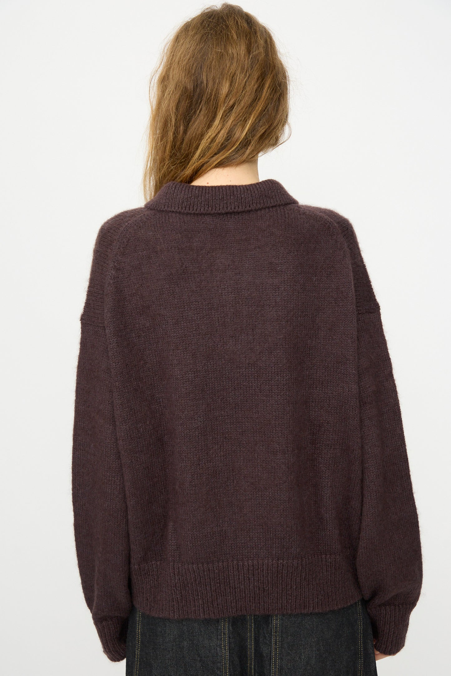 A person with long, light brown hair is seen from the back against a white background, wearing the Studio Nicholson Brushed Mohair Dune Knit Shirt in Espresso—a loose, long-sleeved mohair sweater.