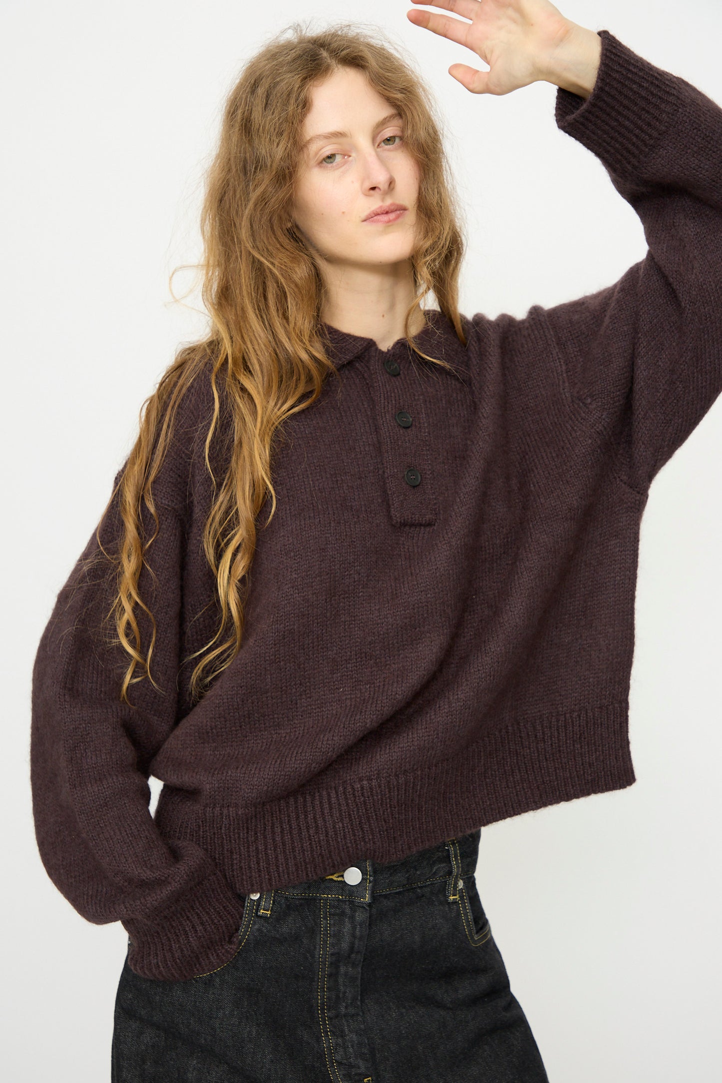 A person with long wavy hair wears the Studio Nicholson Brushed Mohair Dune Knit Shirt in Espresso with black buttons and dark jeans, standing against a plain light background and raising one arm slightly.