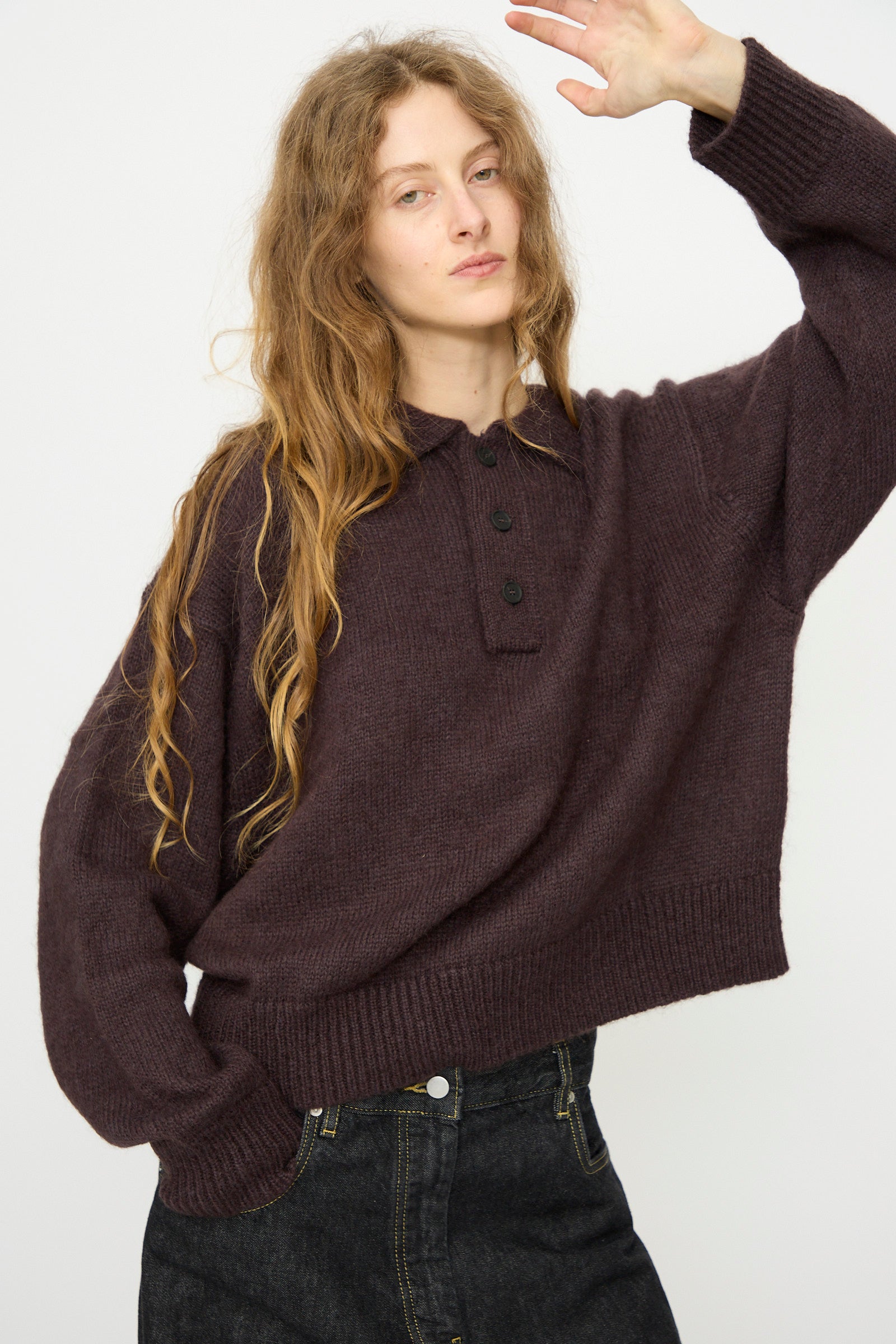 A person with long wavy hair wears the Studio Nicholson Brushed Mohair Dune Knit Shirt in Espresso with black buttons and dark jeans, standing against a plain light background and raising one arm slightly.