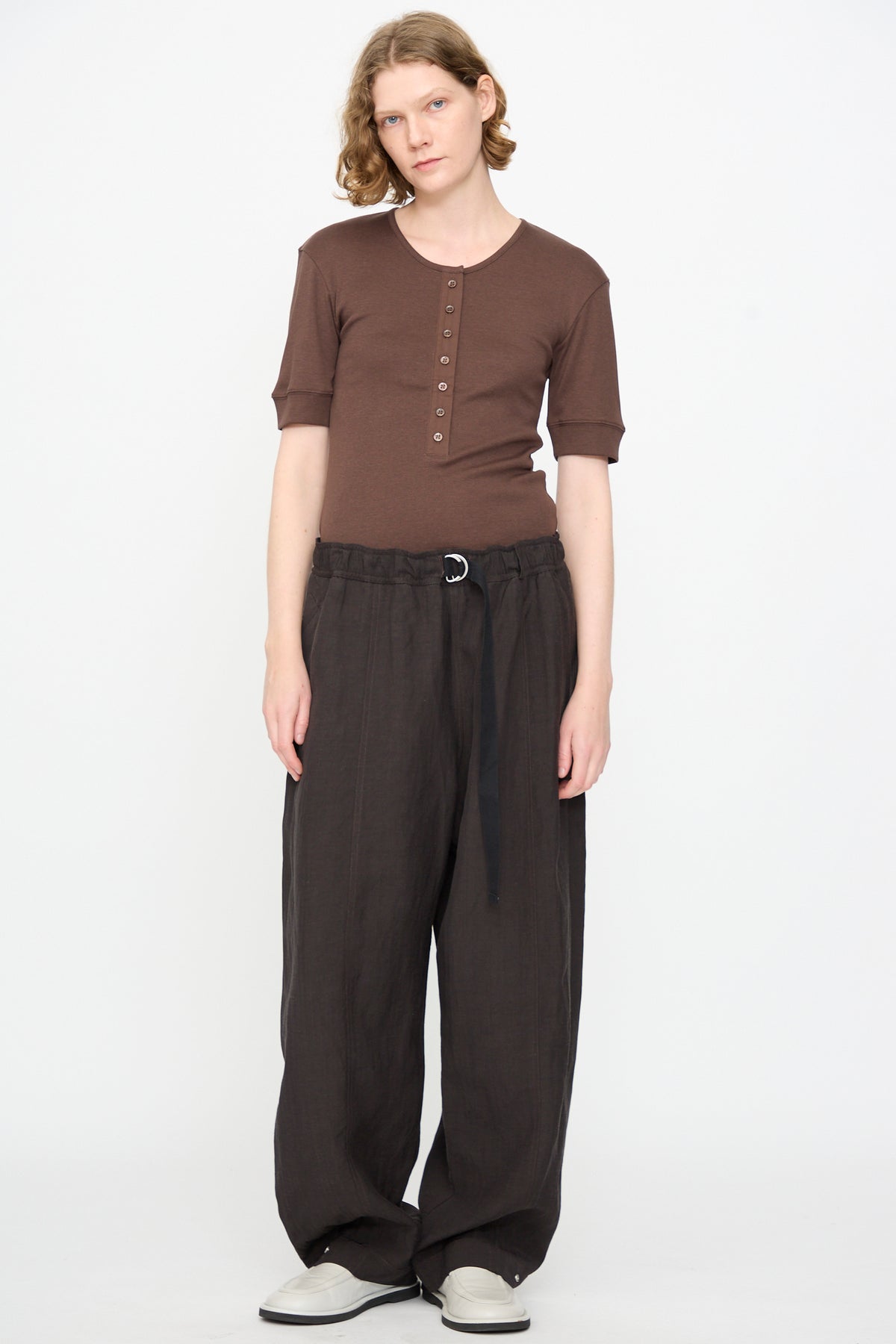 A person stands against a plain white background, wearing a brown short-sleeve henley shirt, Studio Nicholson’s Calvo Pant in Ore—relaxed fit dark brown viscose linen pants with a fabric belt made in Portugal—and white shoes.