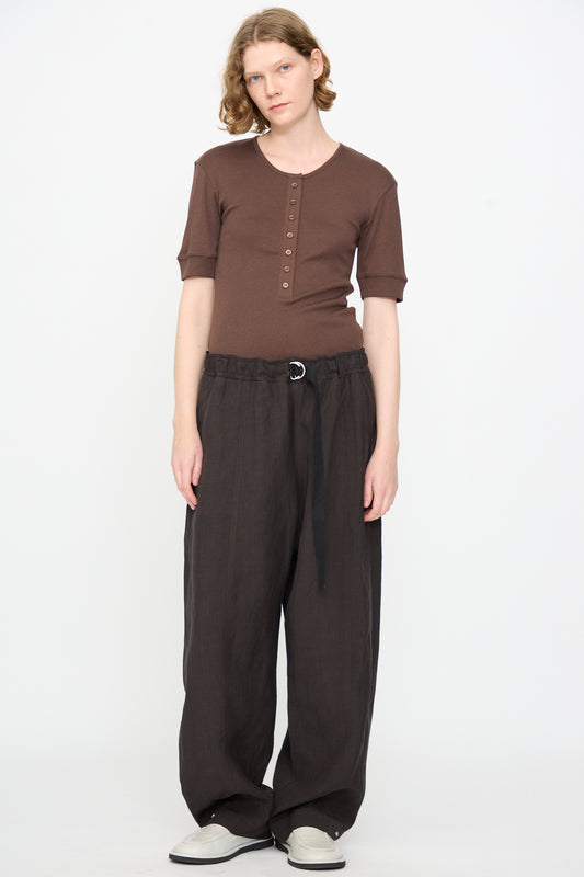 A person stands against a plain white background, wearing a brown short-sleeve henley shirt, Studio Nicholson’s Calvo Pant in Ore—relaxed fit dark brown viscose linen pants with a fabric belt made in Portugal—and white shoes.