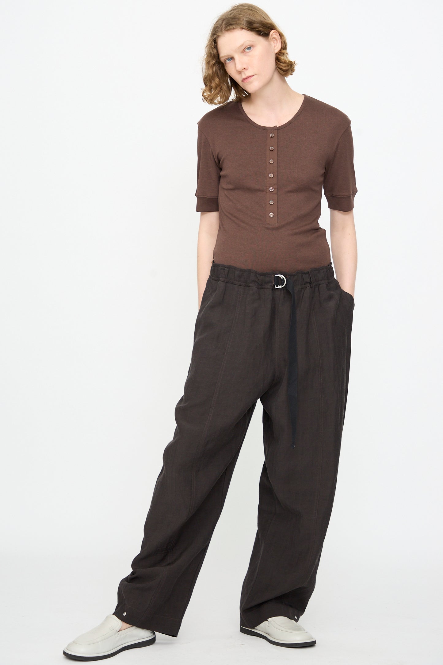 A person wearing a brown short-sleeve henley shirt and Studio Nicholson’s Calvo Pant in Ore—a loose, dark brown viscose linen trouser with a relaxed fit, belted at the waist—stands with hands in pockets against a plain white background. The look effortlessly combines comfort and style, featuring pants thoughtfully made in Portugal.