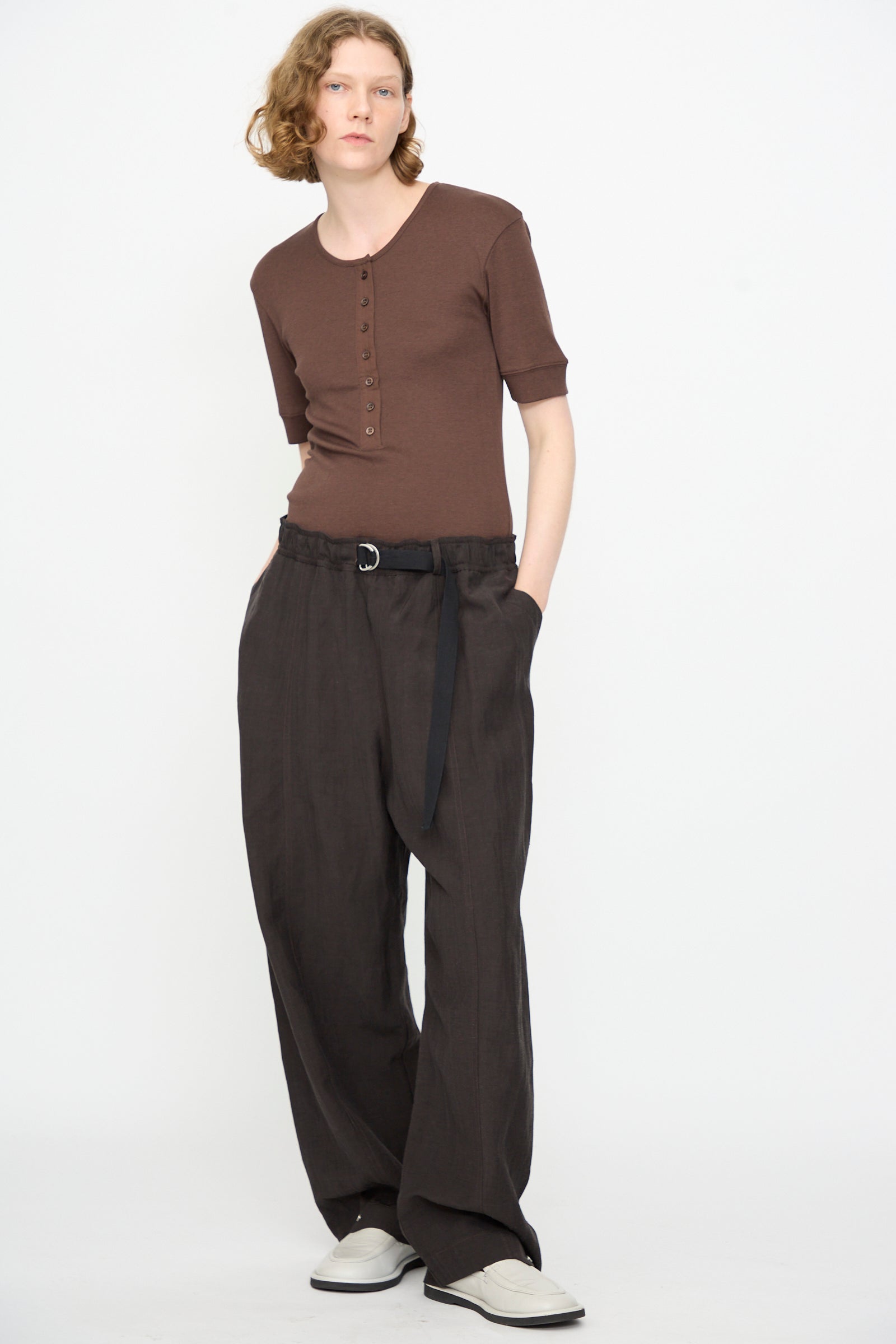 A person is wearing a brown short-sleeve henley shirt, Studio Nicholson Calvo Pant in Ore, styled with a black belt and white shoes, standing against a plain white background.