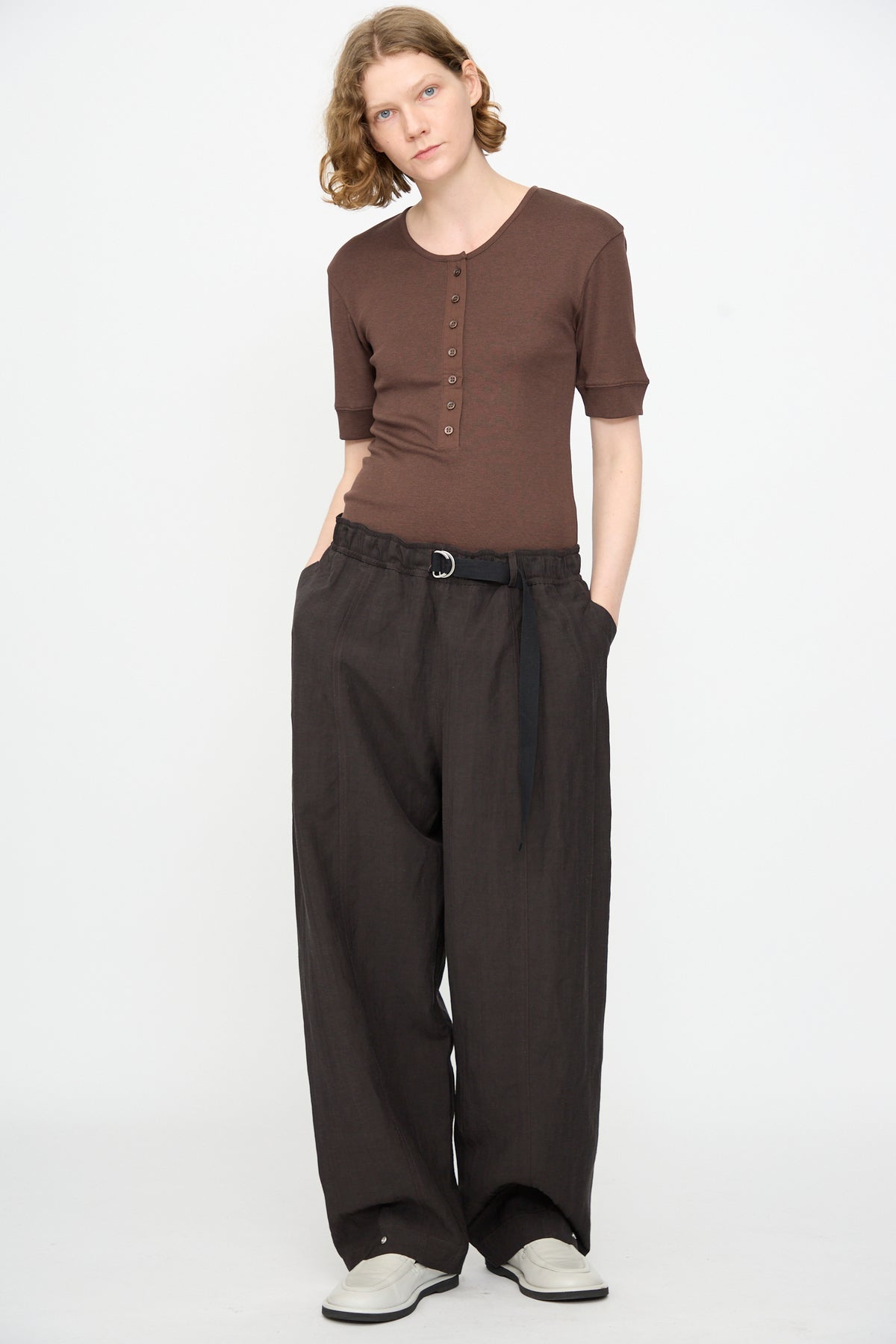 A person with wavy hair wearing a brown buttoned top, the Studio Nicholson Calvo Pant in Ore—a relaxed fit wide-leg viscose linen style—with a black belt and white shoes, standing against a plain white background.