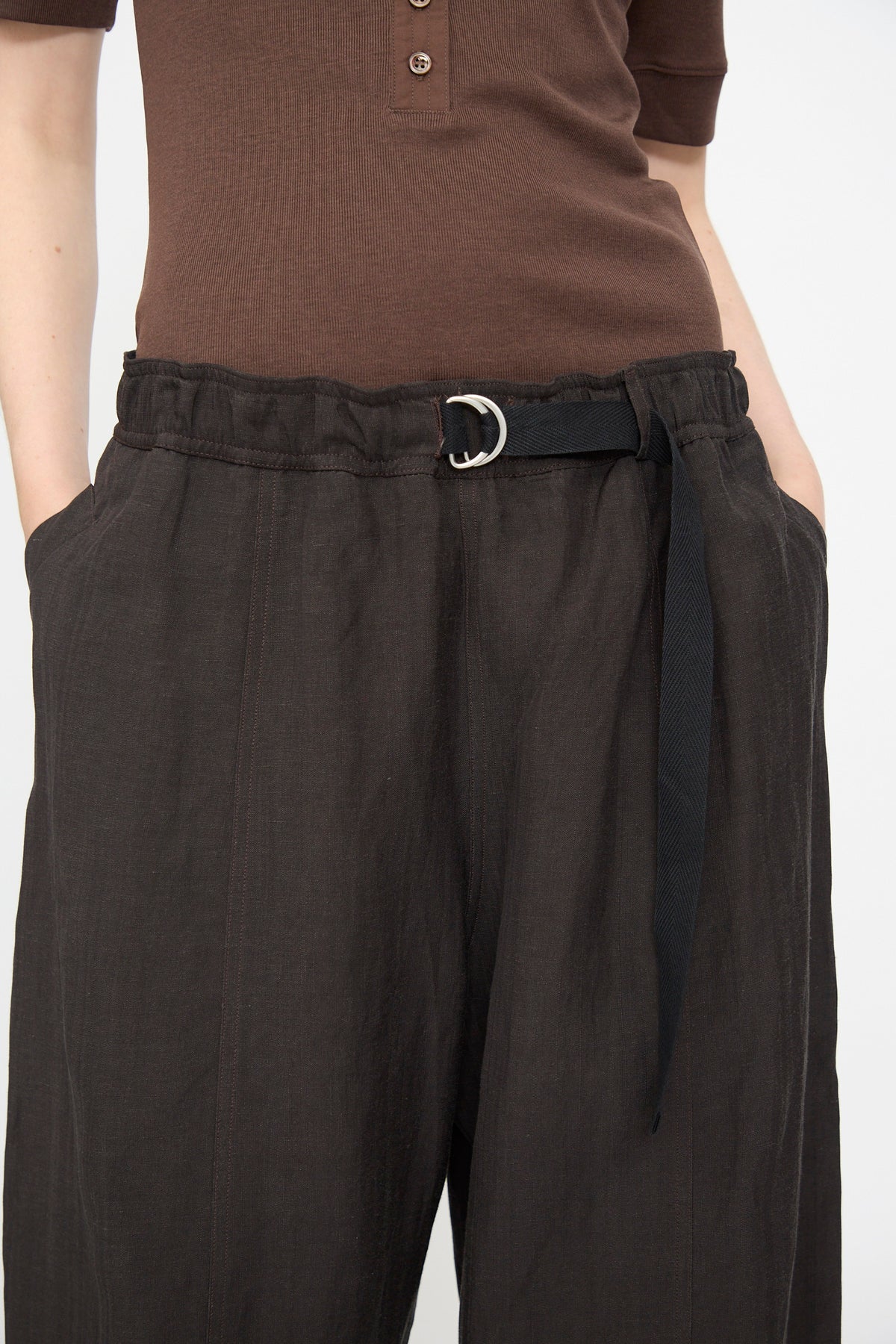A person wears a brown short-sleeve shirt tucked into Studio Nicholson’s Calvo Pant in Ore—dark, relaxed fit viscose linen pants featuring an elastic waistband, black fabric belt, and hands in pockets.
