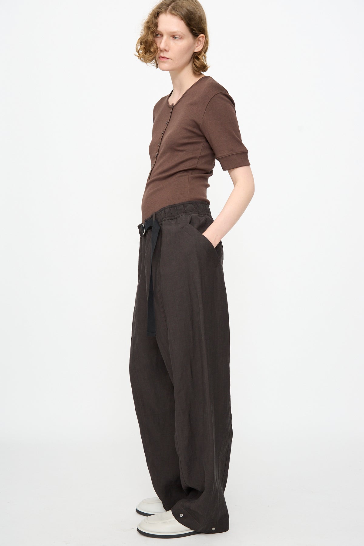 A person stands in profile against a plain background, wearing a brown short-sleeve top, Studio Nicholson’s Calvo Pant in Ore—relaxed fit viscose linen pants with a belt made in Portugal—and white shoes.
