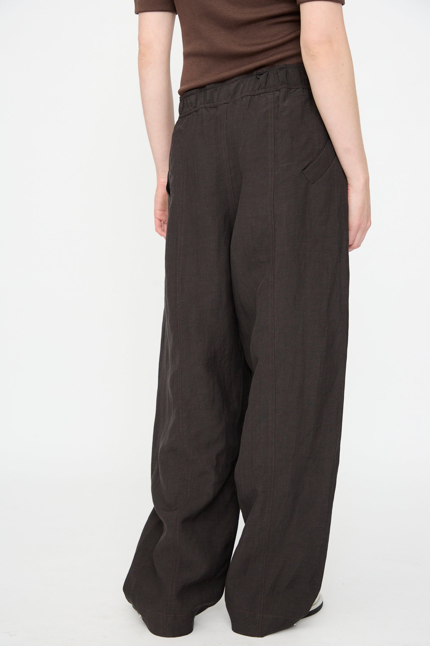 A person stands against a plain white background, shown from the back, wearing the Studio Nicholson Calvo Pant in Ore—loose-fitting, dark brown viscose linen trousers paired with a brown short-sleeve top. The relaxed fit of the pants lends an effortless sense of style to the look.