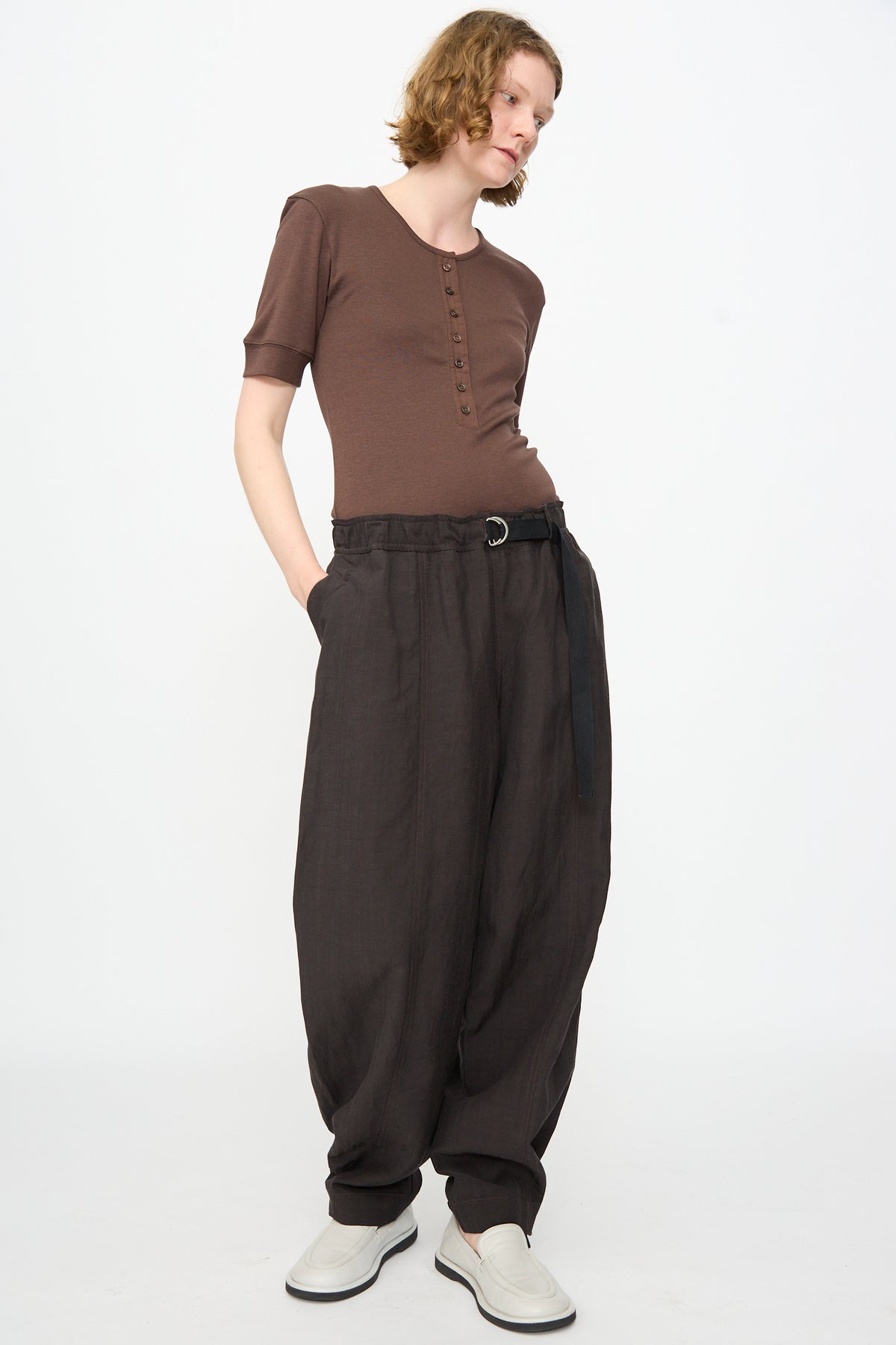 Wearing the Studio Nicholson Calvo Pant in Ore, this person styles the relaxed fit trousers with a brown short-sleeve buttoned top and a belt, complemented by light-colored shoes, standing against a plain white background.