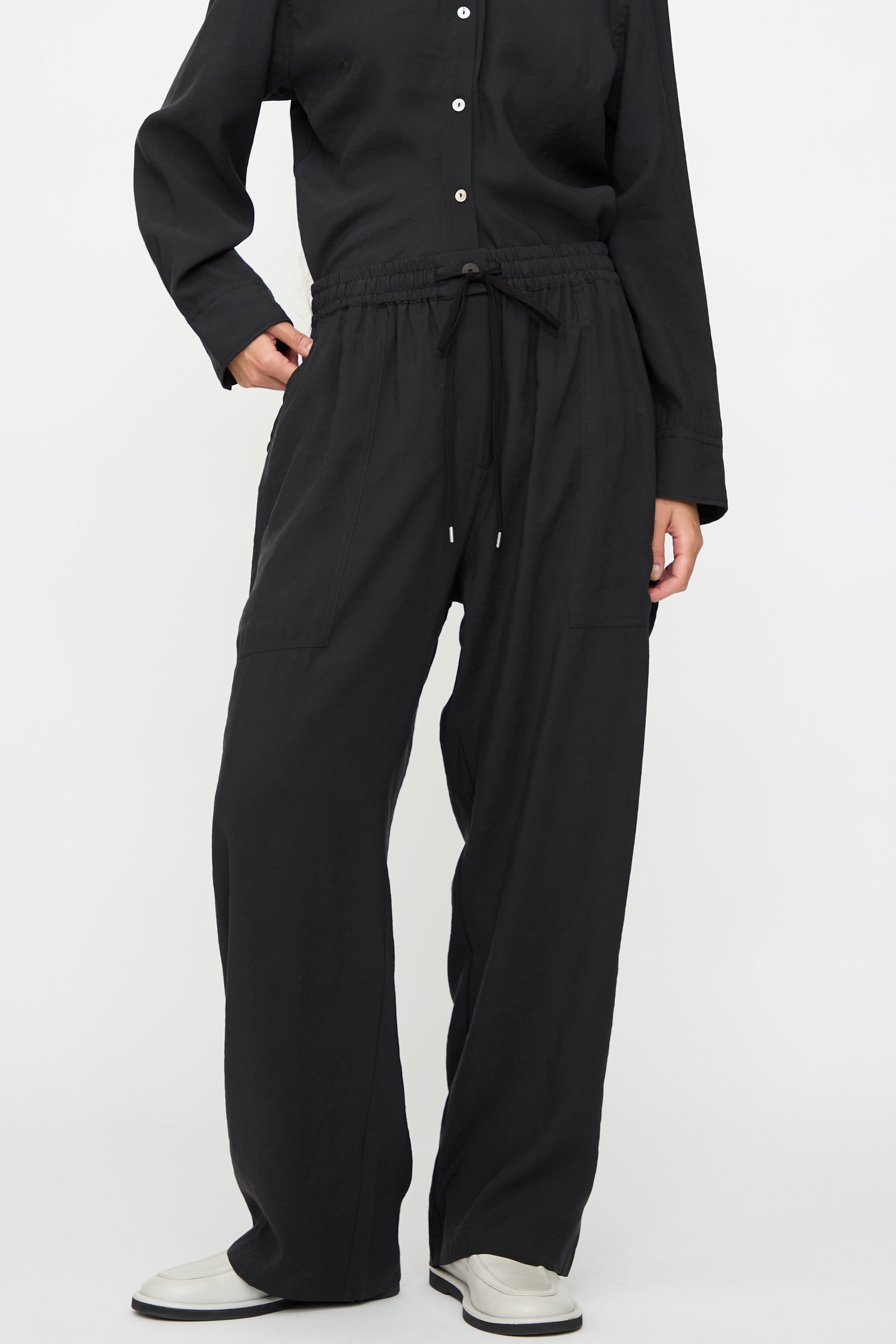 A person wears a long-sleeve black button-up shirt, Studio Nicholson Capel Pant in Darkest Navy (relaxed fit, modal blend), and white shoes, standing against a plain white background.