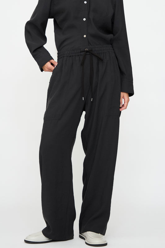 A person wears a long-sleeve black button-up shirt, Studio Nicholson Capel Pant in Darkest Navy (relaxed fit, modal blend), and white shoes, standing against a plain white background.