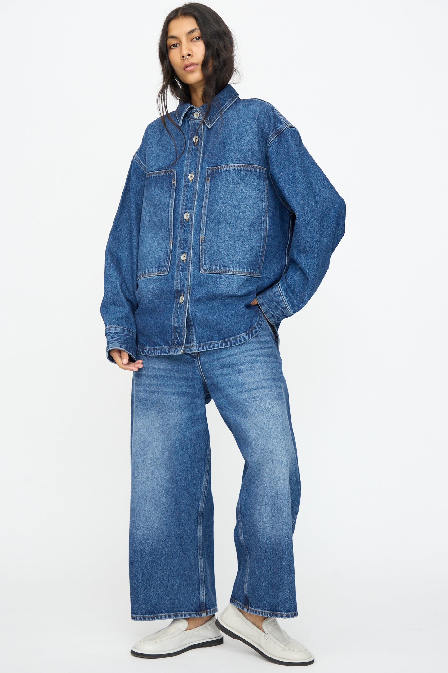 A person stands against a plain white background wearing Studio Nicholson's Chalco Jean in Worn Wash Indigo, styled with a matching oversized denim jacket with large front pockets and white slip-on shoes.
