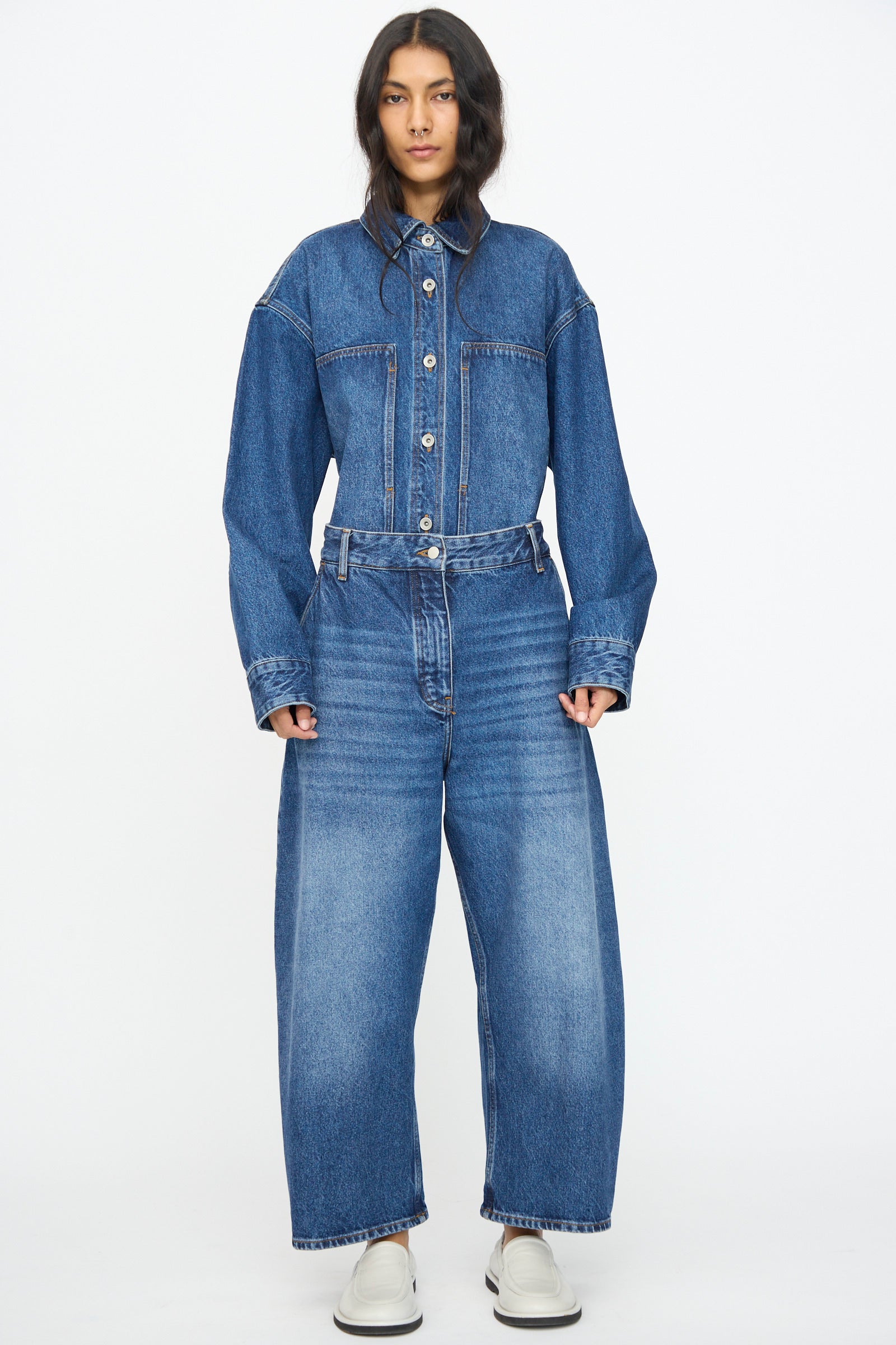 A person wears the Studio Nicholson Chalco Jean in Worn Wash Indigo, styled with a tucked oversized denim shirt, denim belt, and white slip-on shoes, standing against a plain white background.