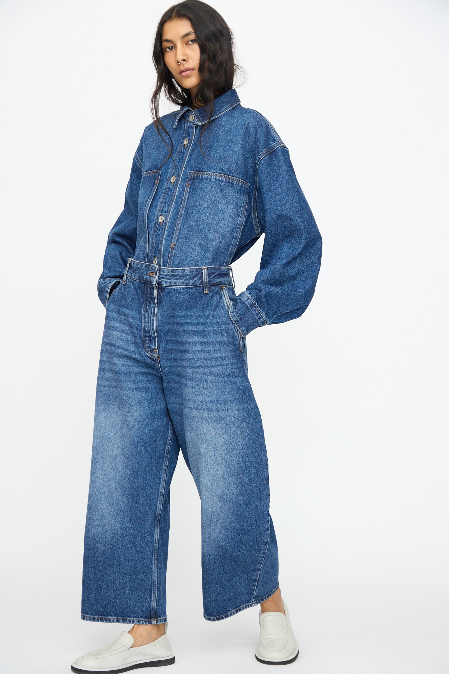 A person stands with hands in pockets, wearing Studio Nicholson’s Chalco Jean in Worn Wash Indigo and a washed denim button-up shirt, paired with white slip-on shoes against a plain, light background.